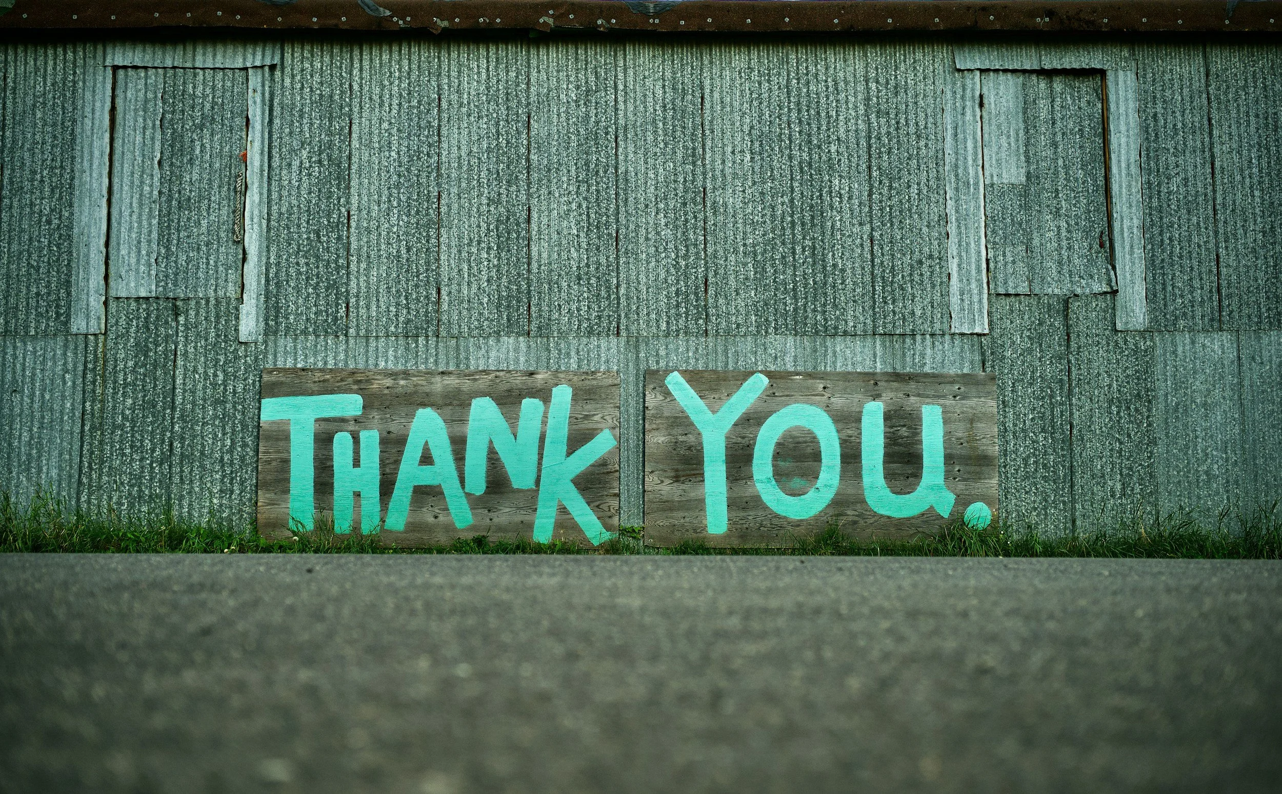 A weathered wooden sign with bright blue letters spelling out "THANK YOU" placed against a rustic, corrugated metal wall on the ground.