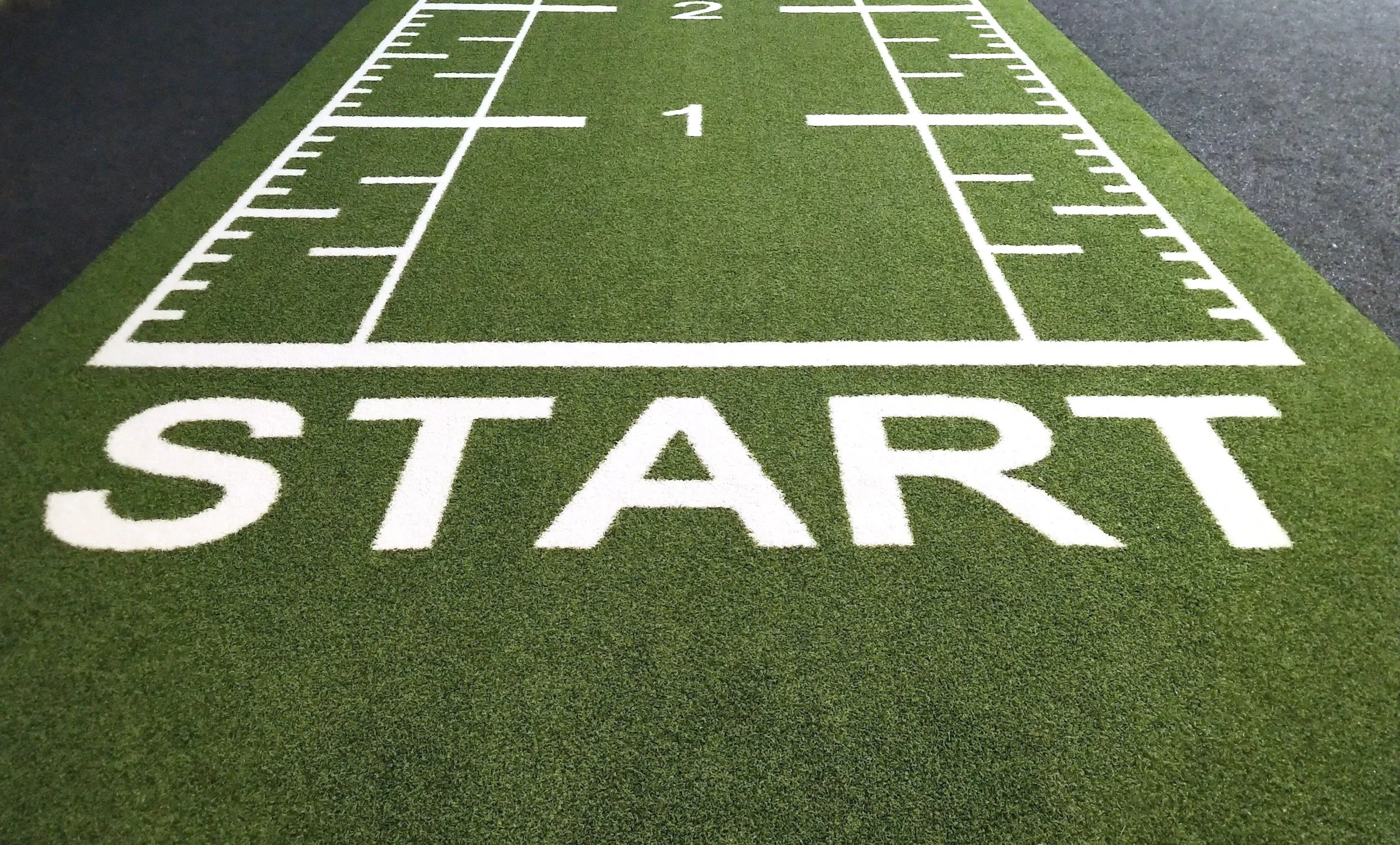 Artificial turf with a white painted running track and the word 'START' at the beginning of the lane, with yard lines and measurements marked along the track.