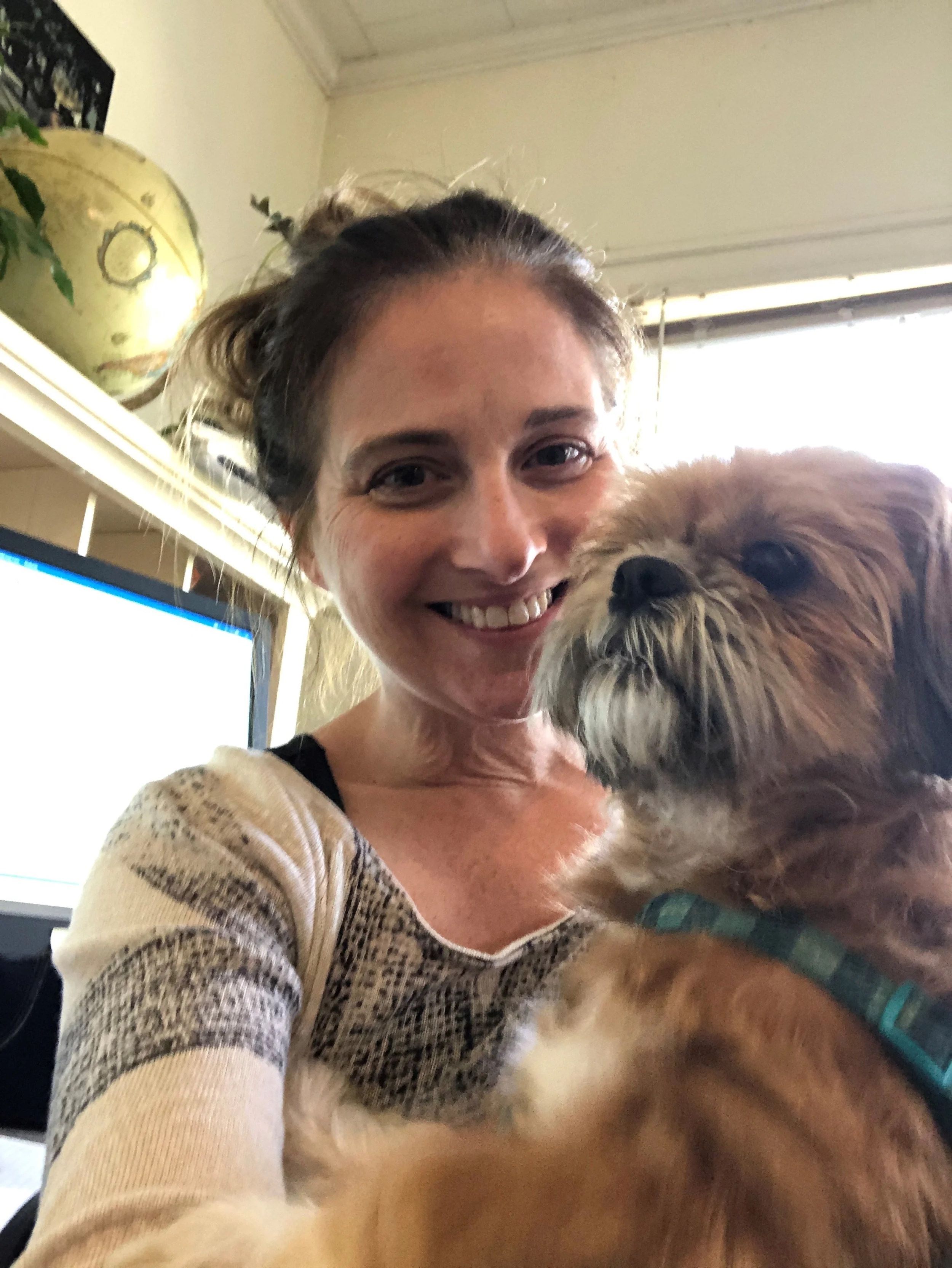 A woman smiling and taking a selfie with her dog, which has long, shaggy fur and is wearing a collar, indoors with a computer monitor and decorative items in the background.