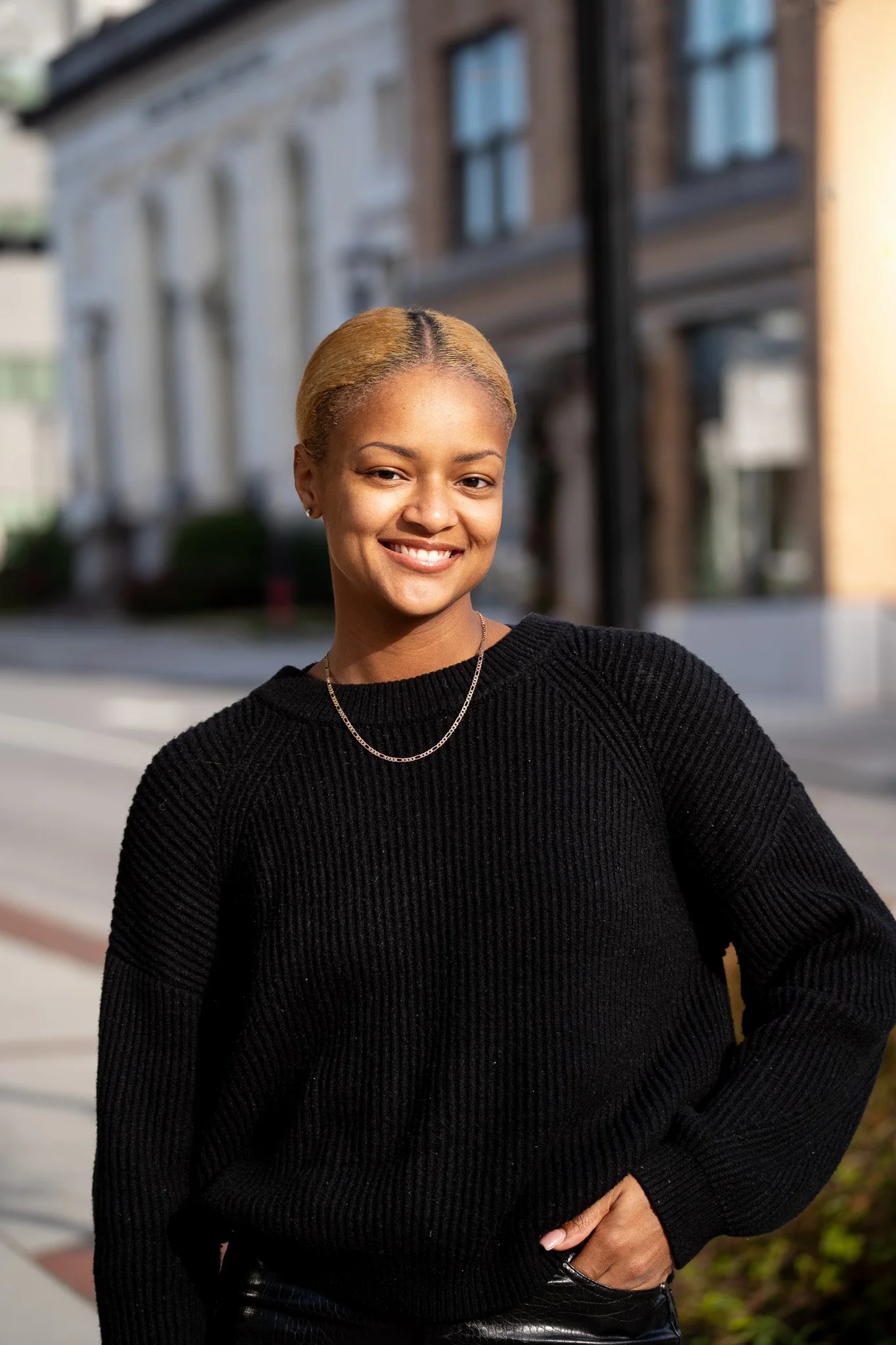 A young woman with blonde hair, wearing a black sweater and necklace, standing outdoors on a city street, smiling at the camera.