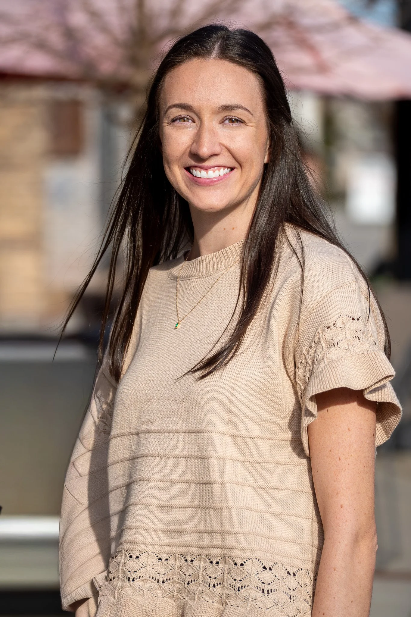 Smiling woman with long dark hair wearing a beige top with lace details on the sleeves and bottom, standing outdoors with buildings and trees in the background during daytime.
