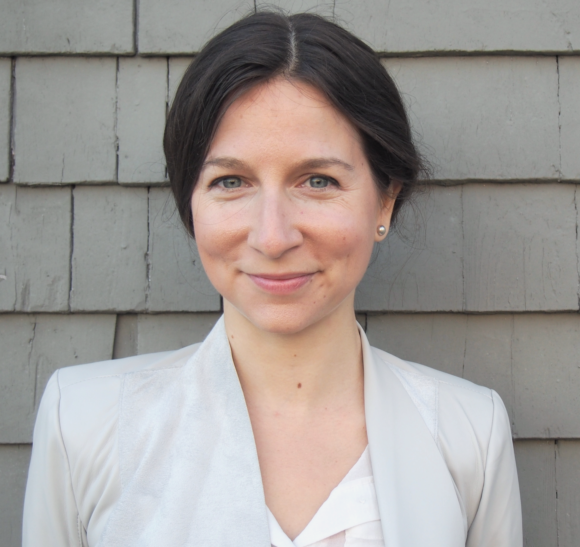 A woman with short dark hair and light skin smiling, wearing a light-colored blazer and shirt, standing in front of a beige brick wall.