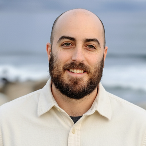 A man with a beard and mustache wearing a blue blazer and a light blue collared shirt, standing outdoors with a blurred background of trees and people.