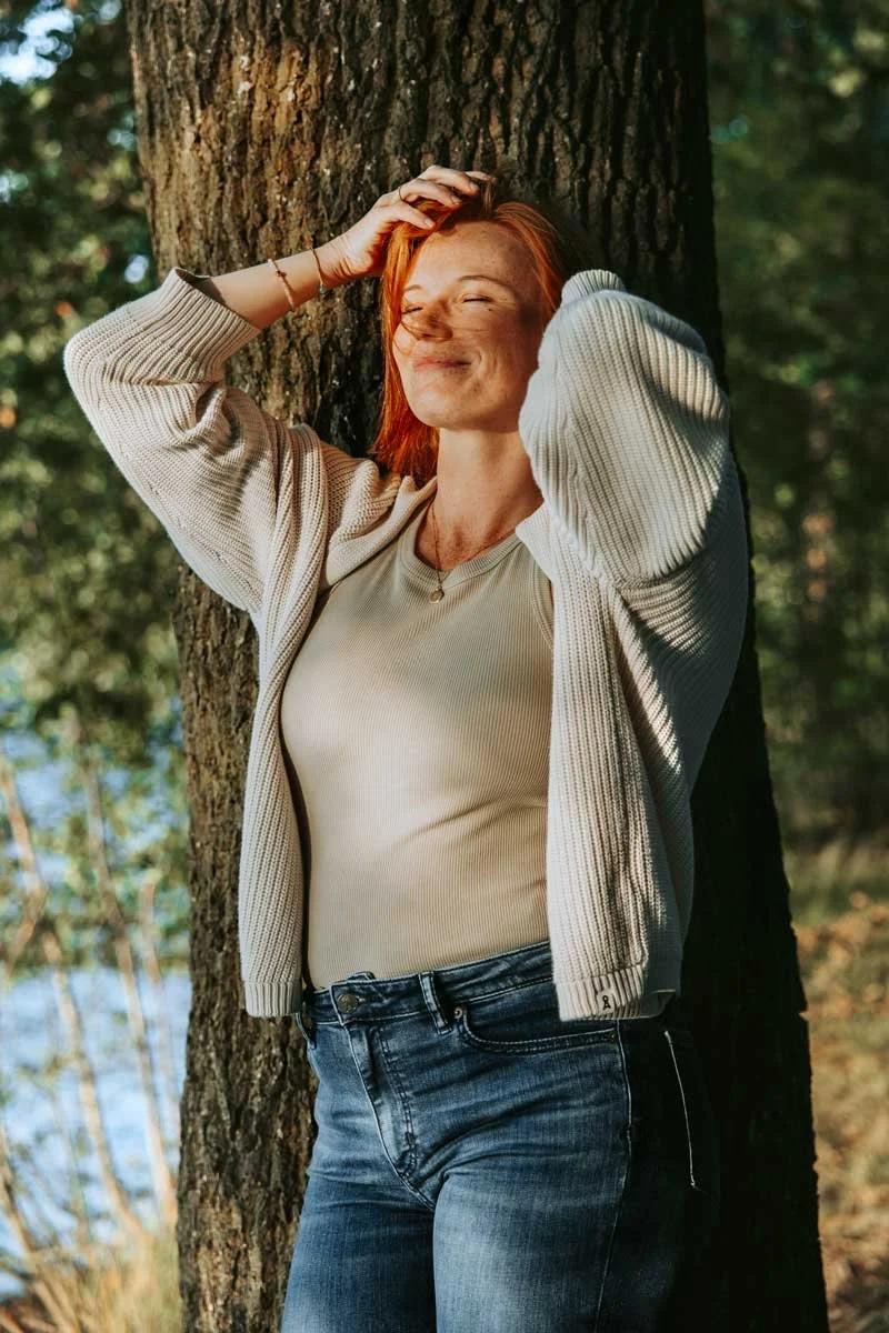 Eine lachende Frau mit roten Haaren lehnt an einem Baum im Wald, die Augen sind fast geschlossen, sie trägt eine beige Jacke und eine cremefarbene Bluse.