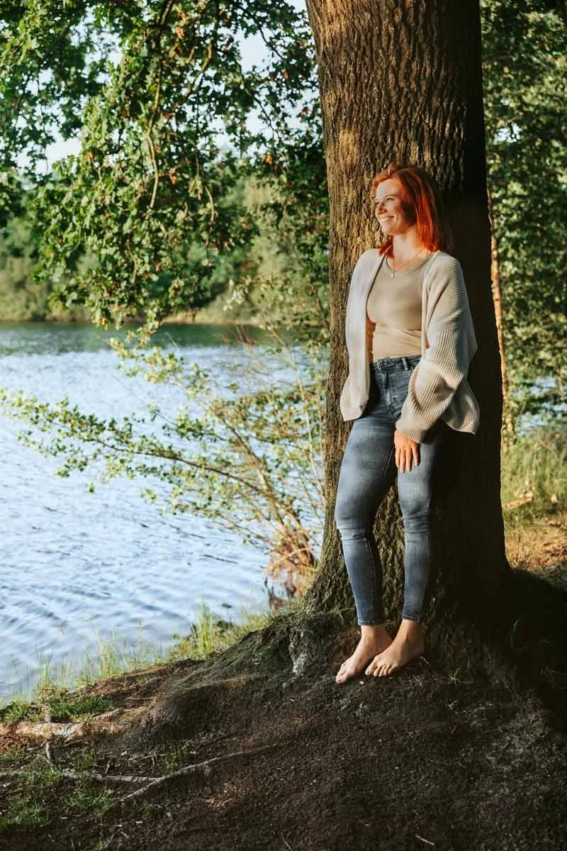 Eine junge Frau mit roten Haaren, legerer Kleidung und barfuß lehnt an einem Baum am Flussufer, lachend, in der Natur bei Sonnenuntergang.