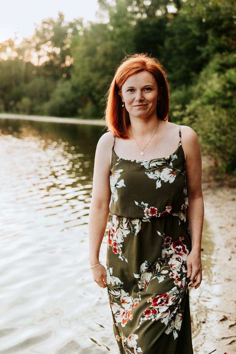 Eine Frau mit roten Haaren in einem grünen, blumigen Kleid steht am Ufer eines Sees mit Bäumen im Hintergrund bei Sonnenuntergang.