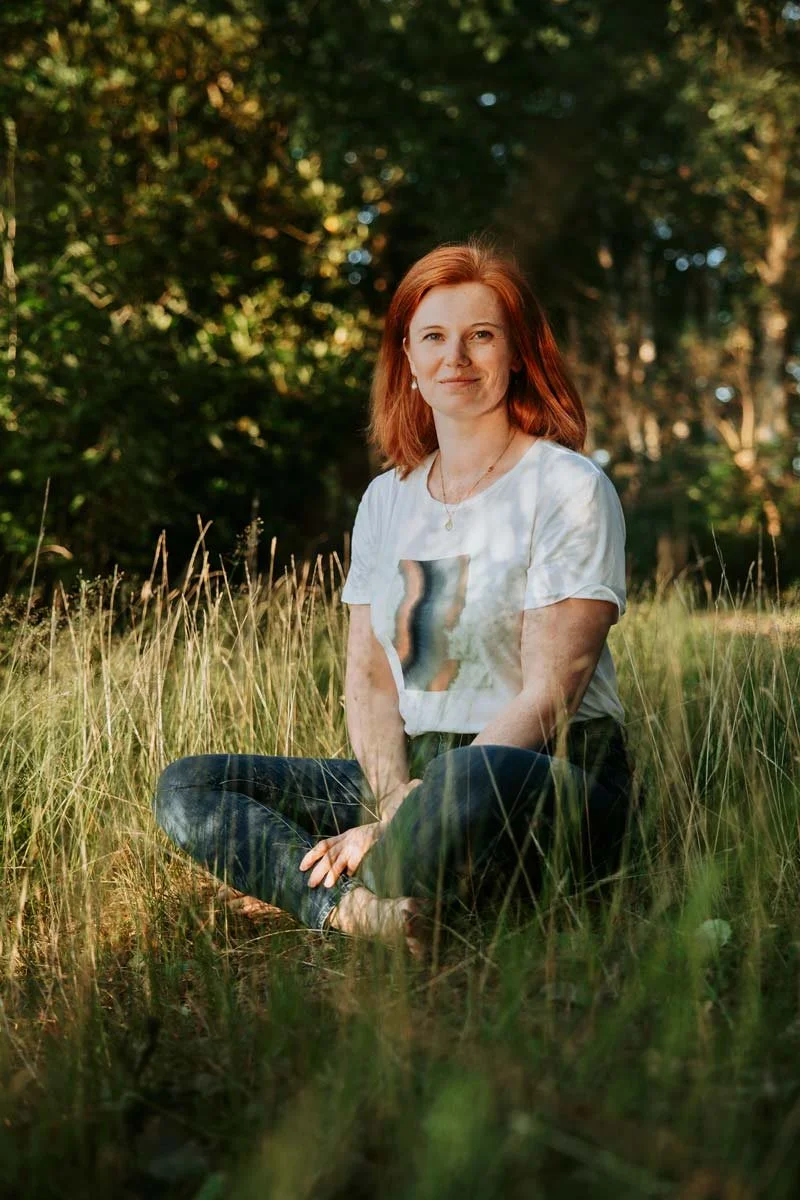 Eine Frau mit roten Haaren sitzt entspannt im hohen Gras in einem Wald, umgeben von Bäumen, im Sonnenschein, lächelnd in die Kamera schauend.