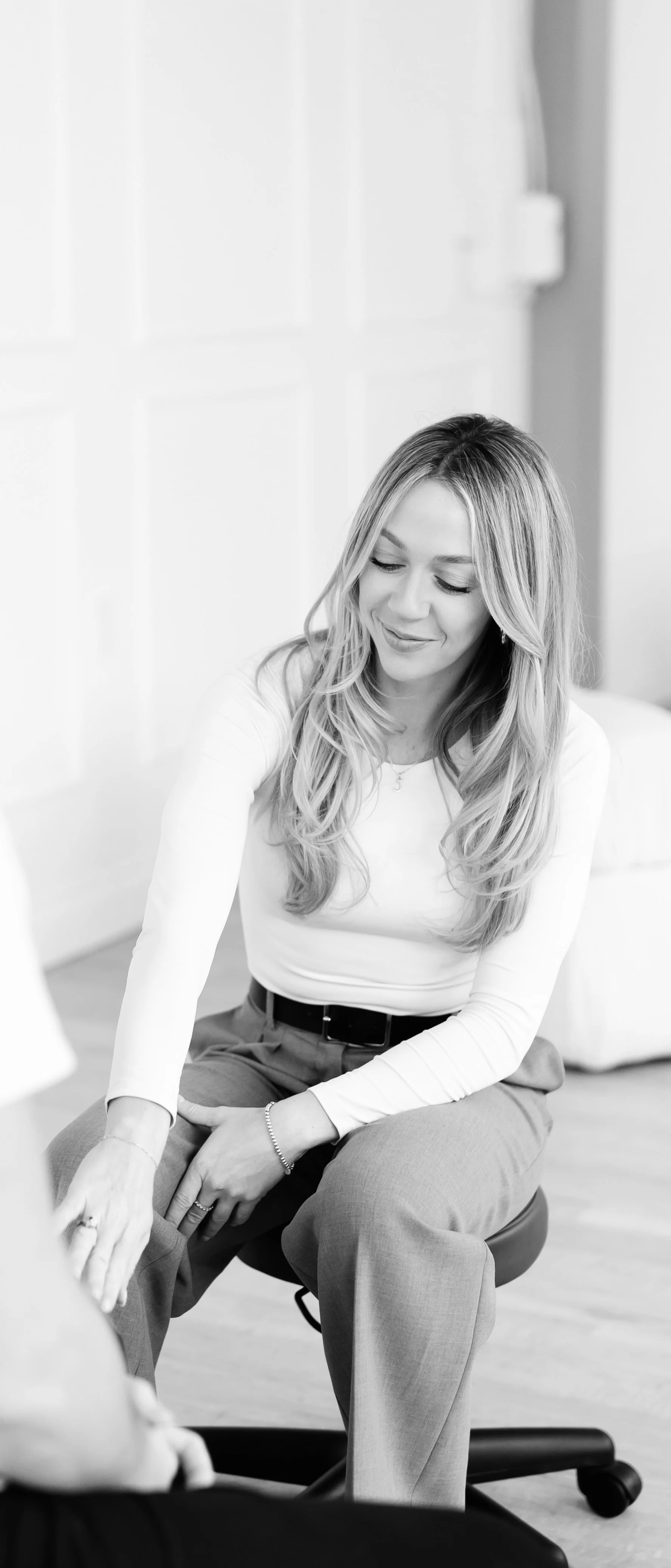 A woman with long wavy hair, wearing a white long-sleeve top and gray pants, sitting on a chair in a bright room, smiling softly and engaging in a conversation.