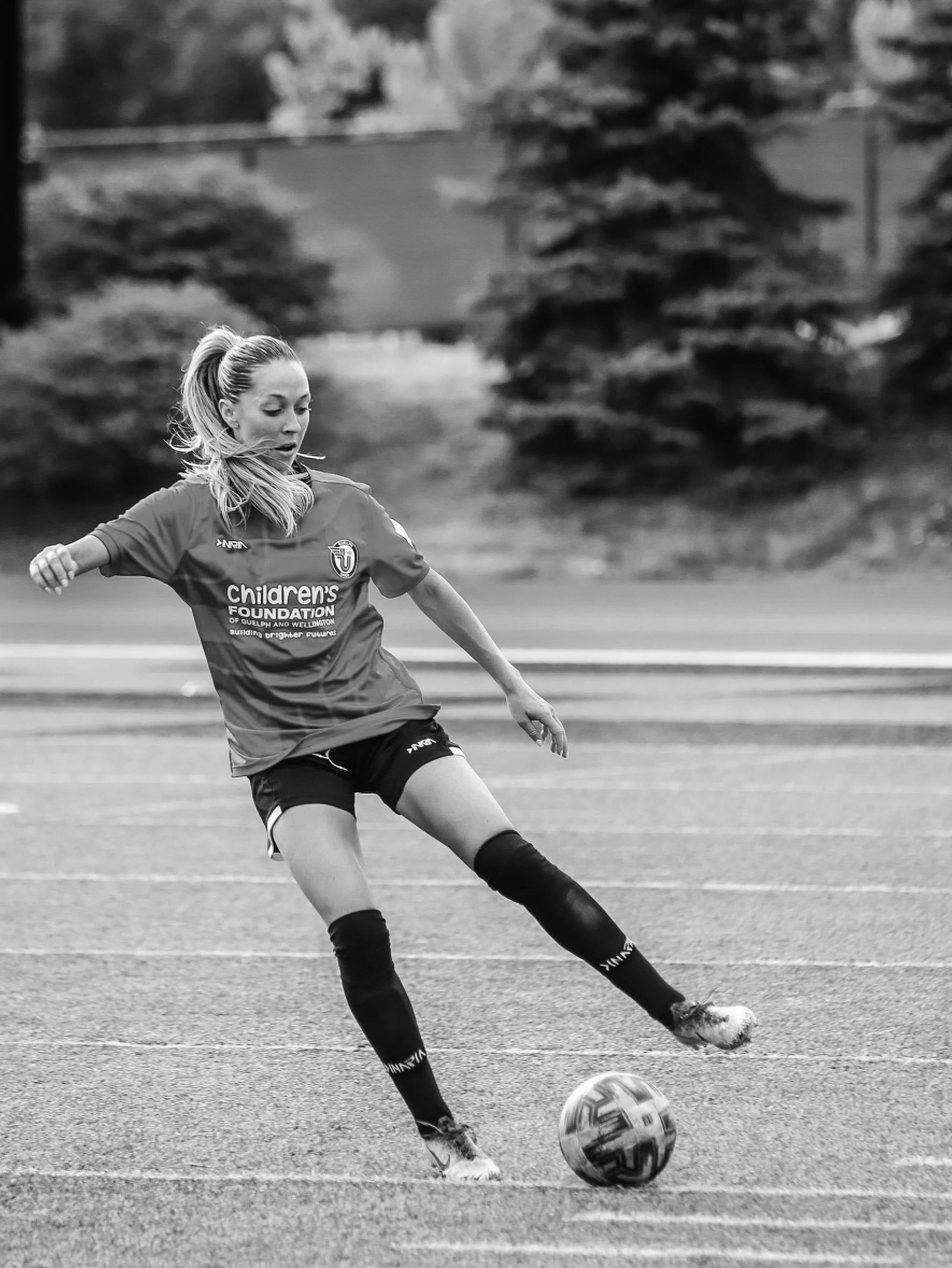 Black and white photo of a young woman kicking a soccer ball on a field, wearing sports attire with a logo and text on her shirt, with trees and a fence in the background.