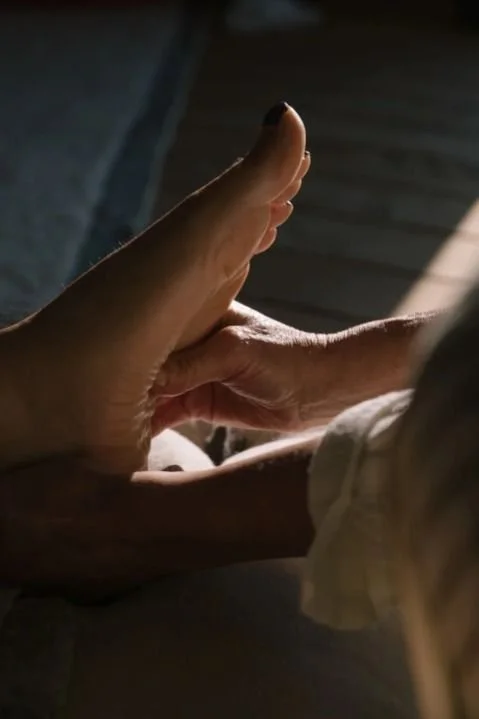 Une main touche le pied d'une autre personne pour lui masser la voûte plantaire dans une ambiance tamisée.