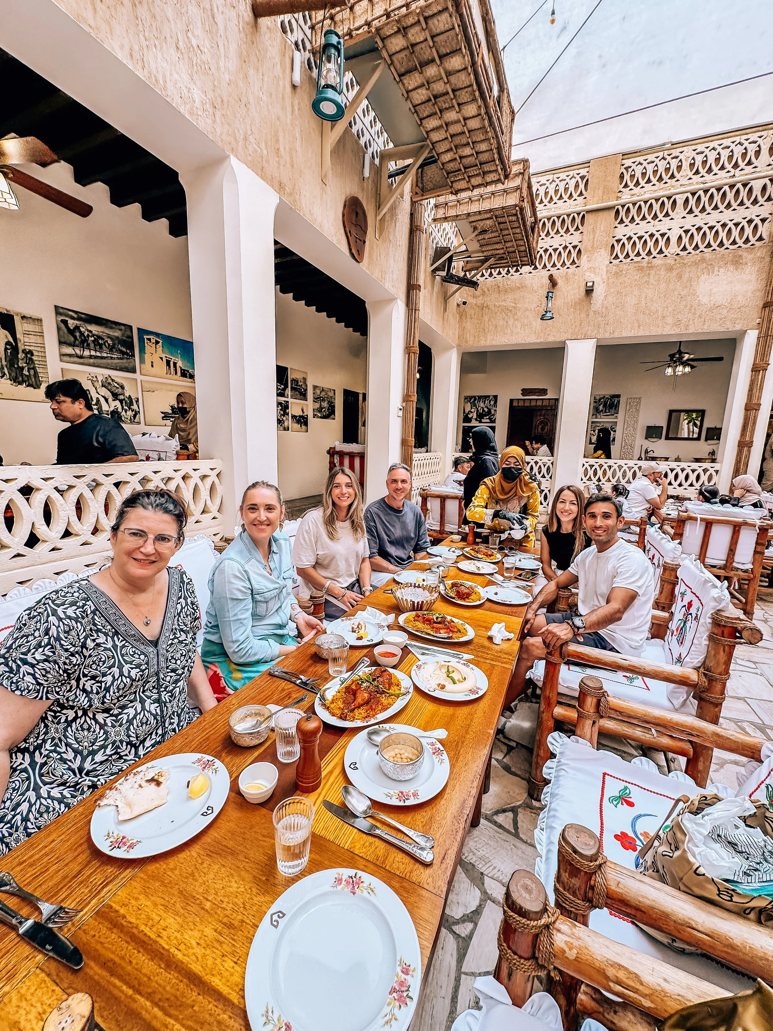 People dining outdoors at a long wooden table with plates of food, drinks, and cutlery in a rustic restaurant with open-air decor and artwork on the walls.