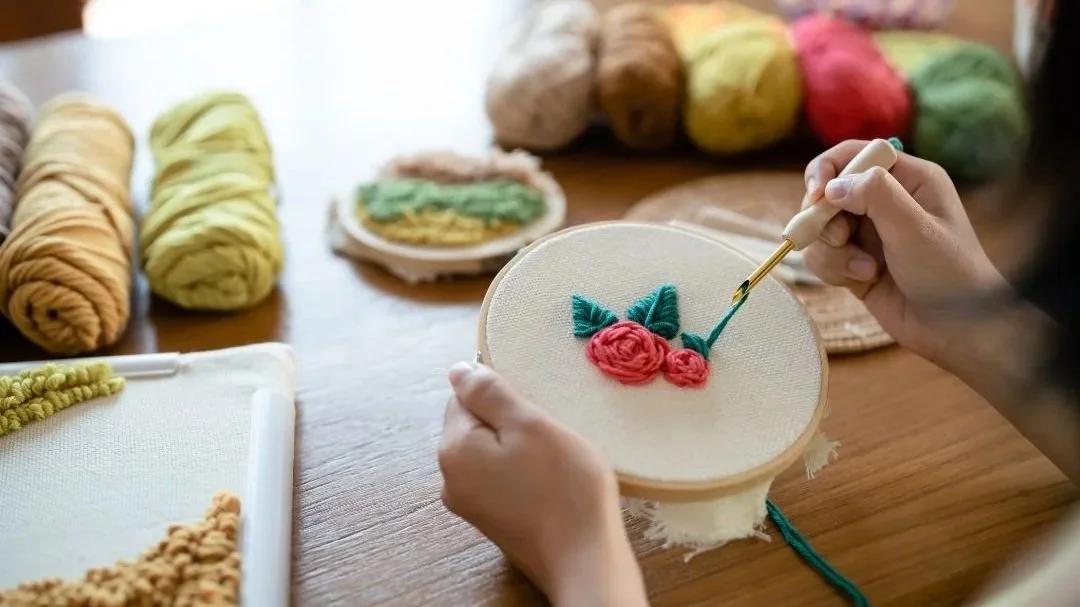 A person creating an embroidery design of a cherry on fabric stretched in an embroidery hoop, with colorful yarns and embroidery supplies in the background.
