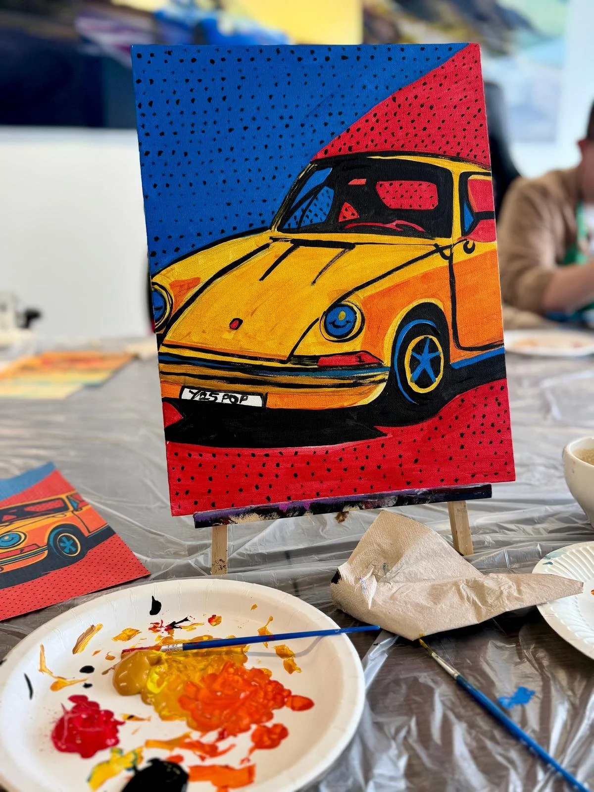 A colorful painting of a yellow vintage car with blue accents, set against a background of blue and red with black dots, displayed on a small easel on a table with painter's supplies.
