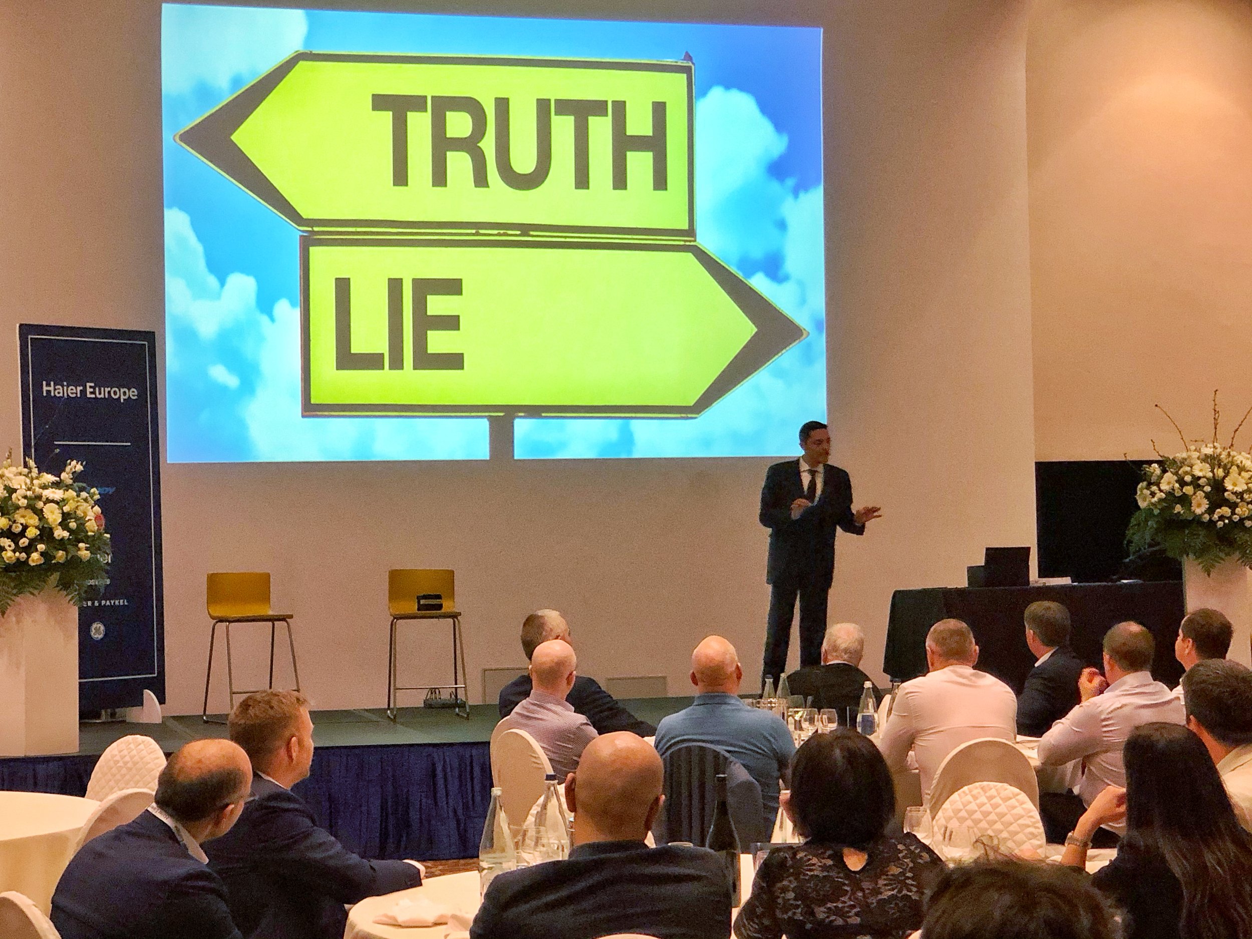 Performer in a suit presenting a mentalism show, with a large screen displaying a road sign pointing left labelled 'TRUTH' and right labelled 'LIE'. Audience members are seated at round tables.