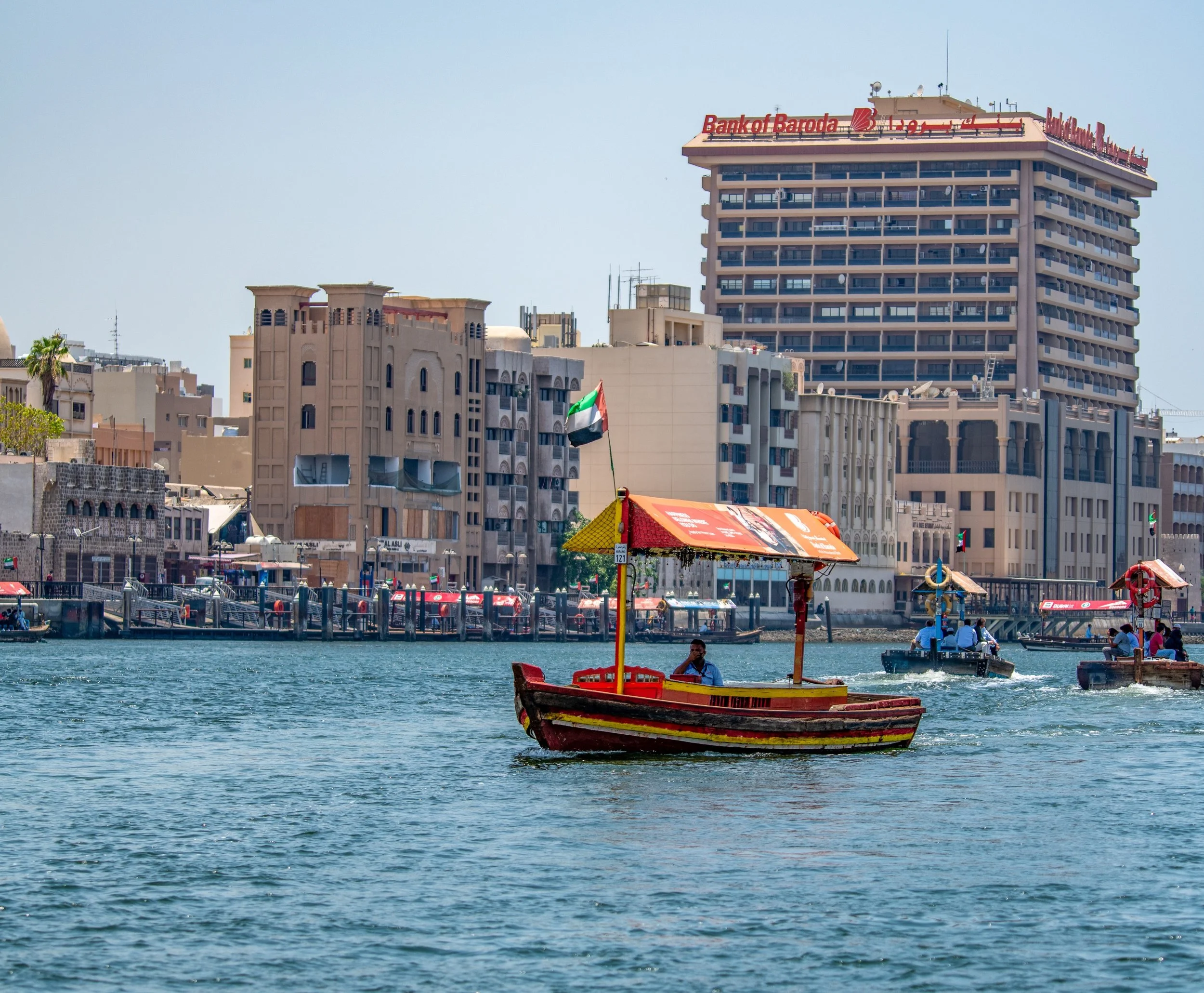 Small boat with a red canopy and a flag sailing on a body of water with a cityscape in the background, including high-rise buildings and waterfront structures.