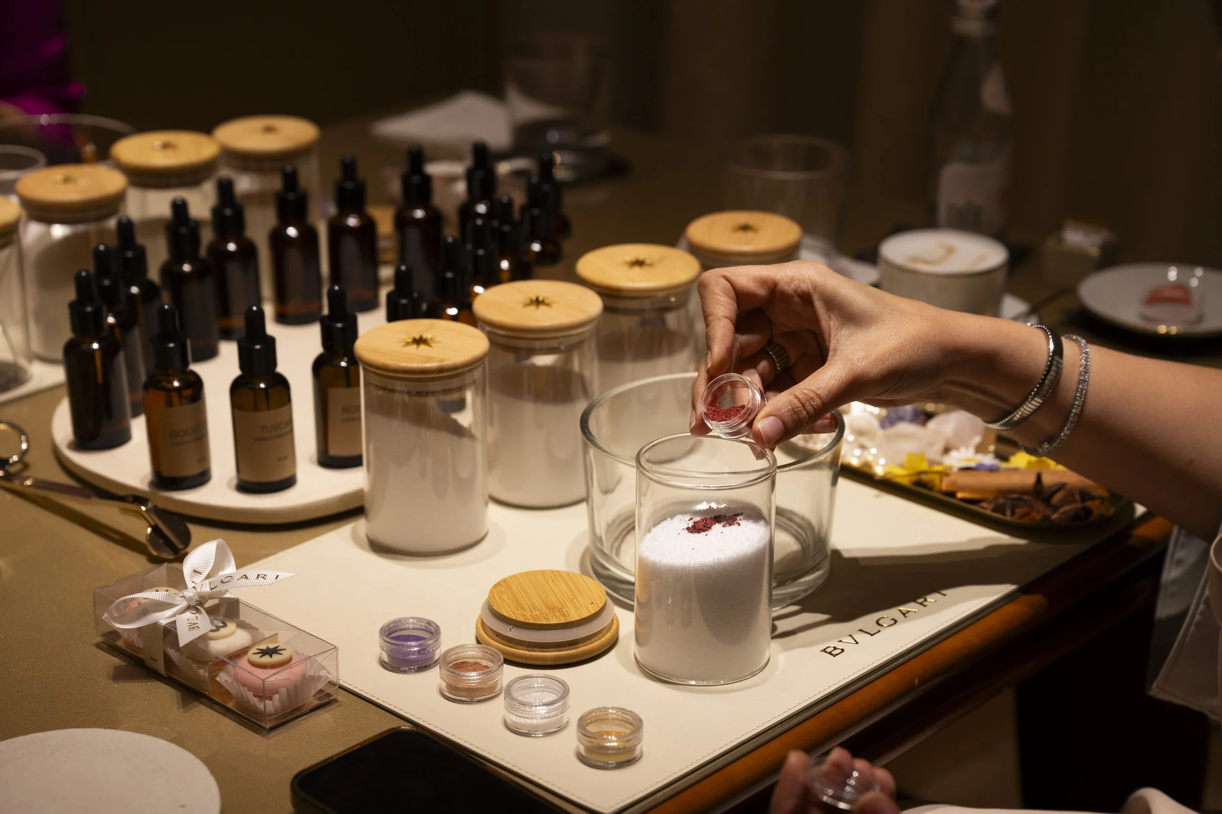 A person is pouring a pink powder into a glass container filled with white granular substance, surrounded by various jars and bottles on a table with a 'BVLGARI' logo.