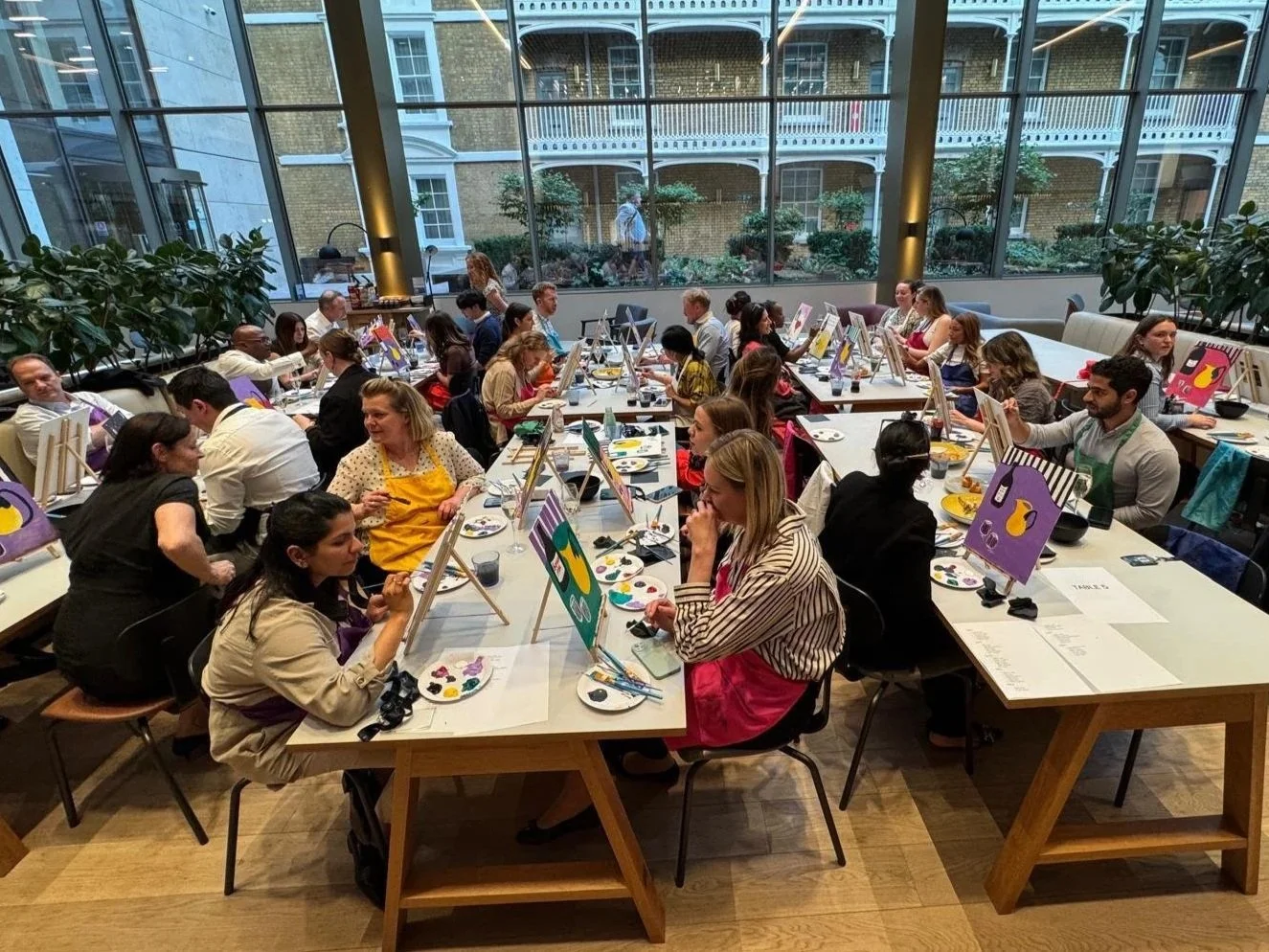 People participating in a group painting class in a bright, spacious room with large windows, each working on individual paintings.