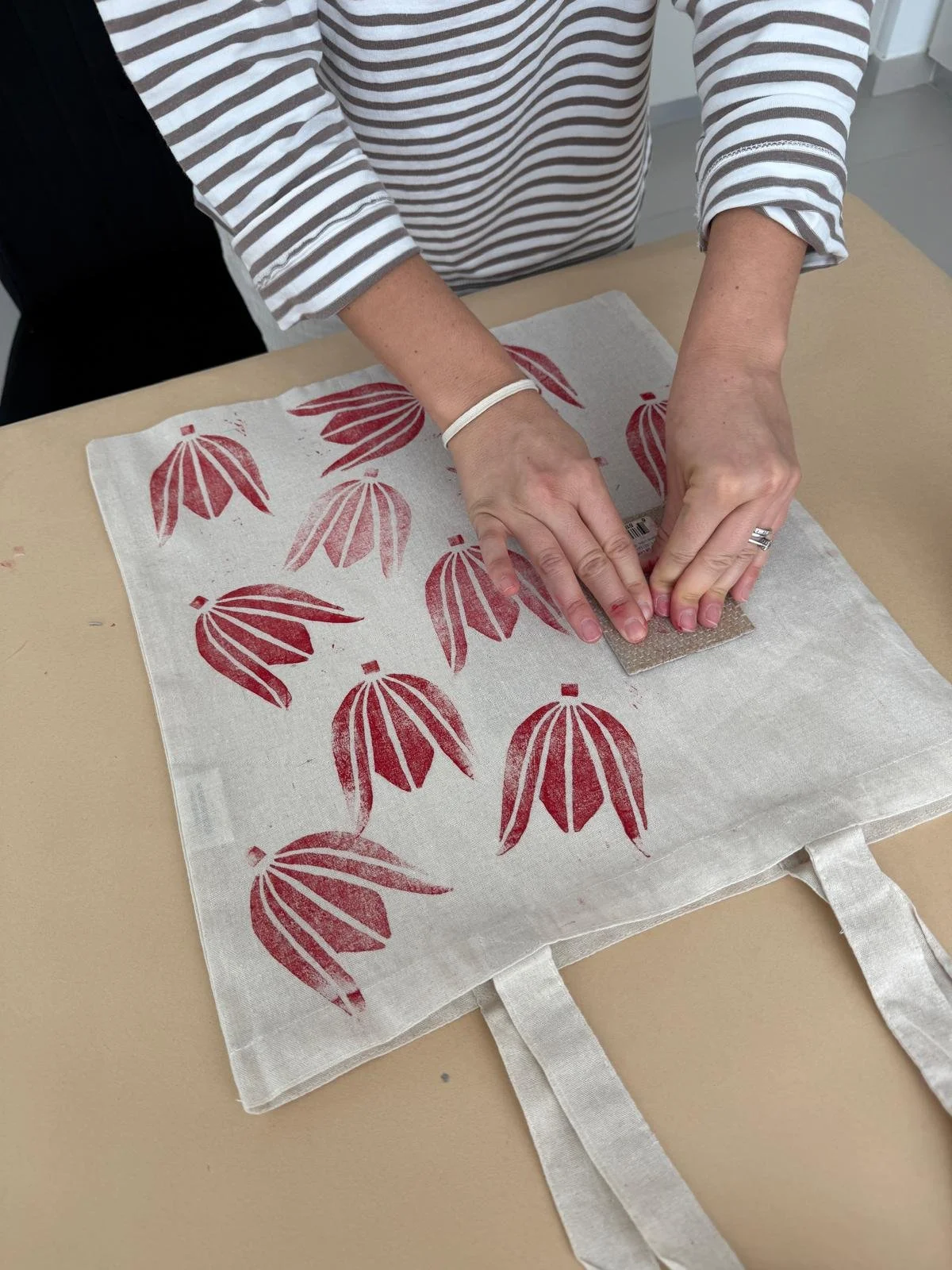 Person using a sanding block to etch or paint a design on a fabric tote bag with a red floral pattern, on a beige table.