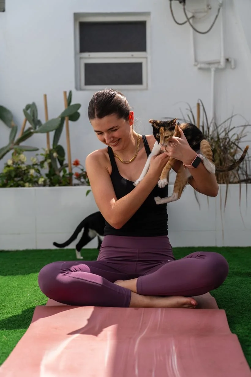 A woman practicing yoga outdoors on a pink mat, holding a calico cat, both smiling, with a green artificial grass surface, plants, and a white wall with a window in the background.