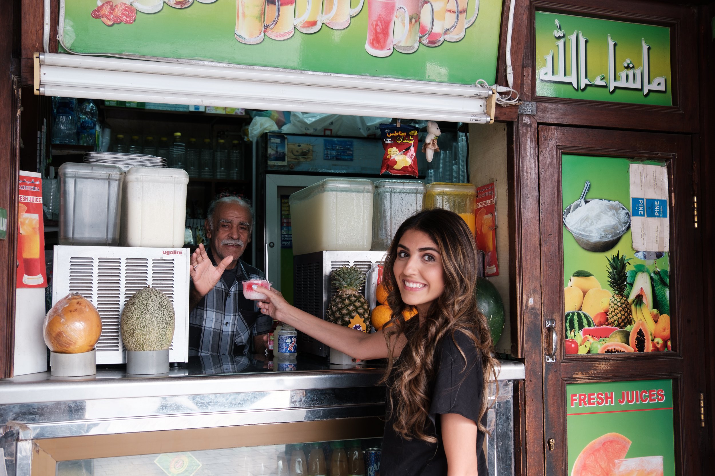 A young woman with long, wavy hair smiling and handing money to an older male vendor at a juice stand. The stand displays various fruits like pineapple and melon, and signs advertising fresh juices and fruit drinks.