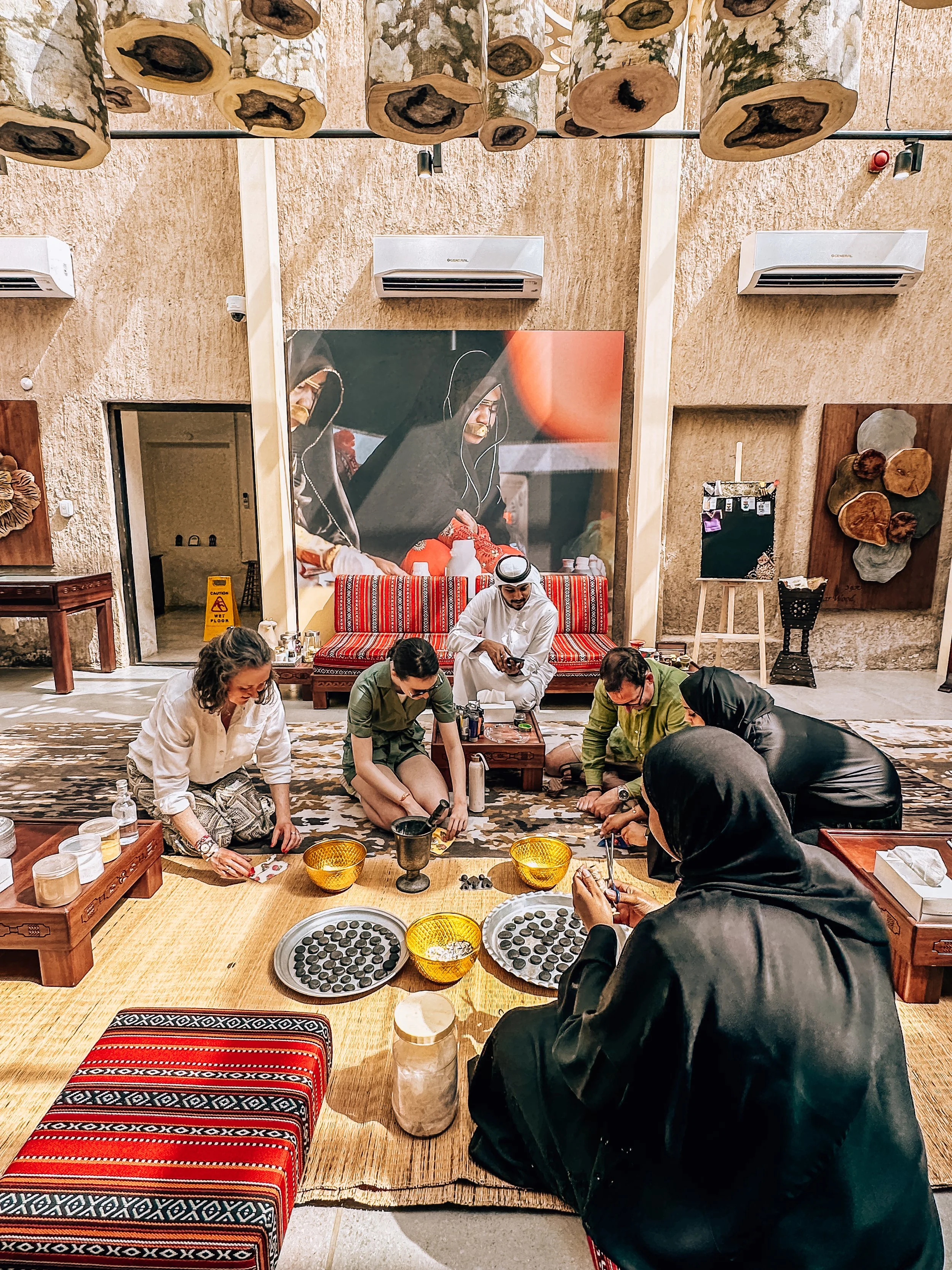 Group of people sitting on the floor around trays of black candies in a room with cultural decor, including wall art and wooden accents.