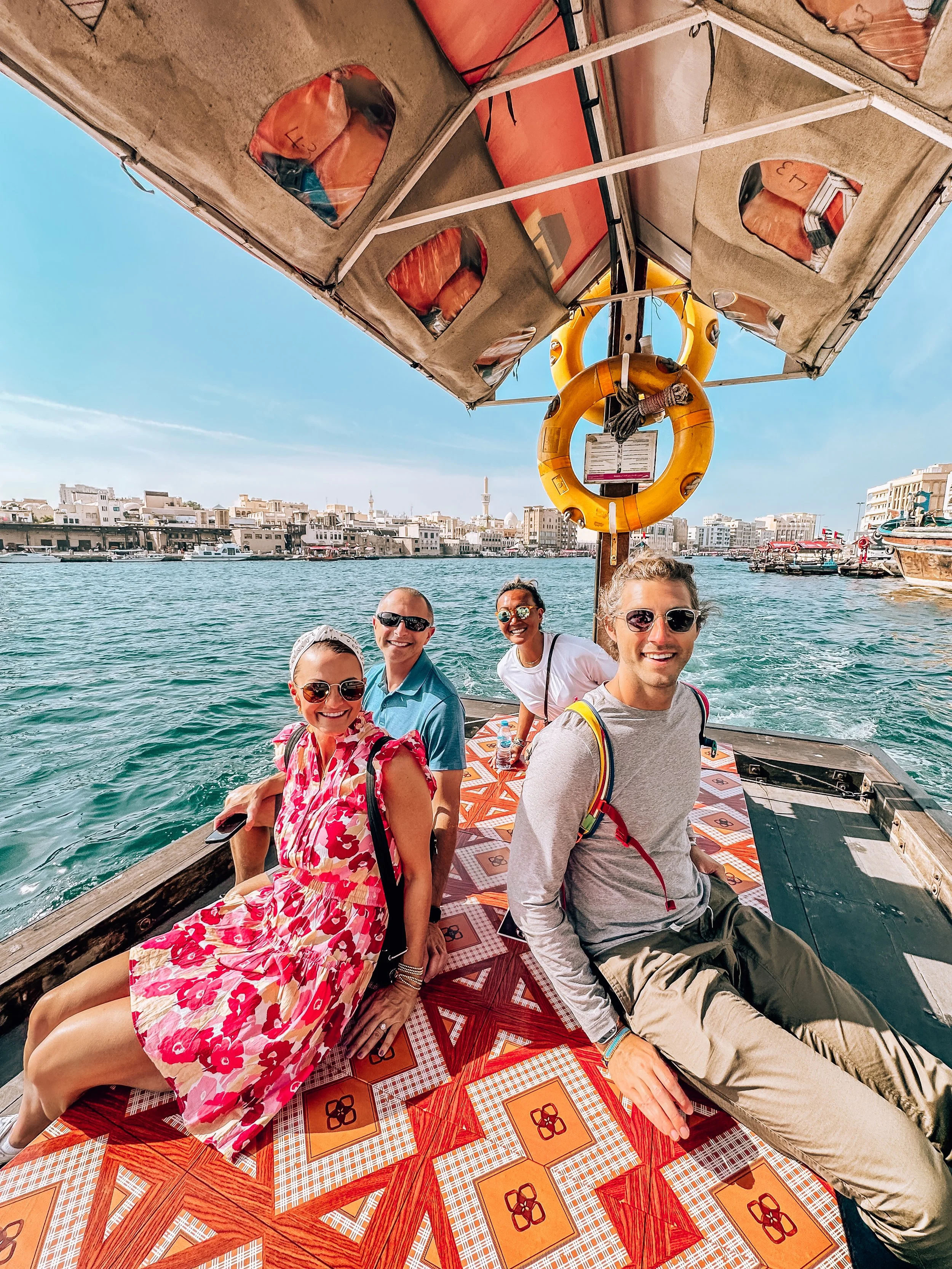 People sitting on a boat in a harbor, with buildings in the background and a clear blue sky.