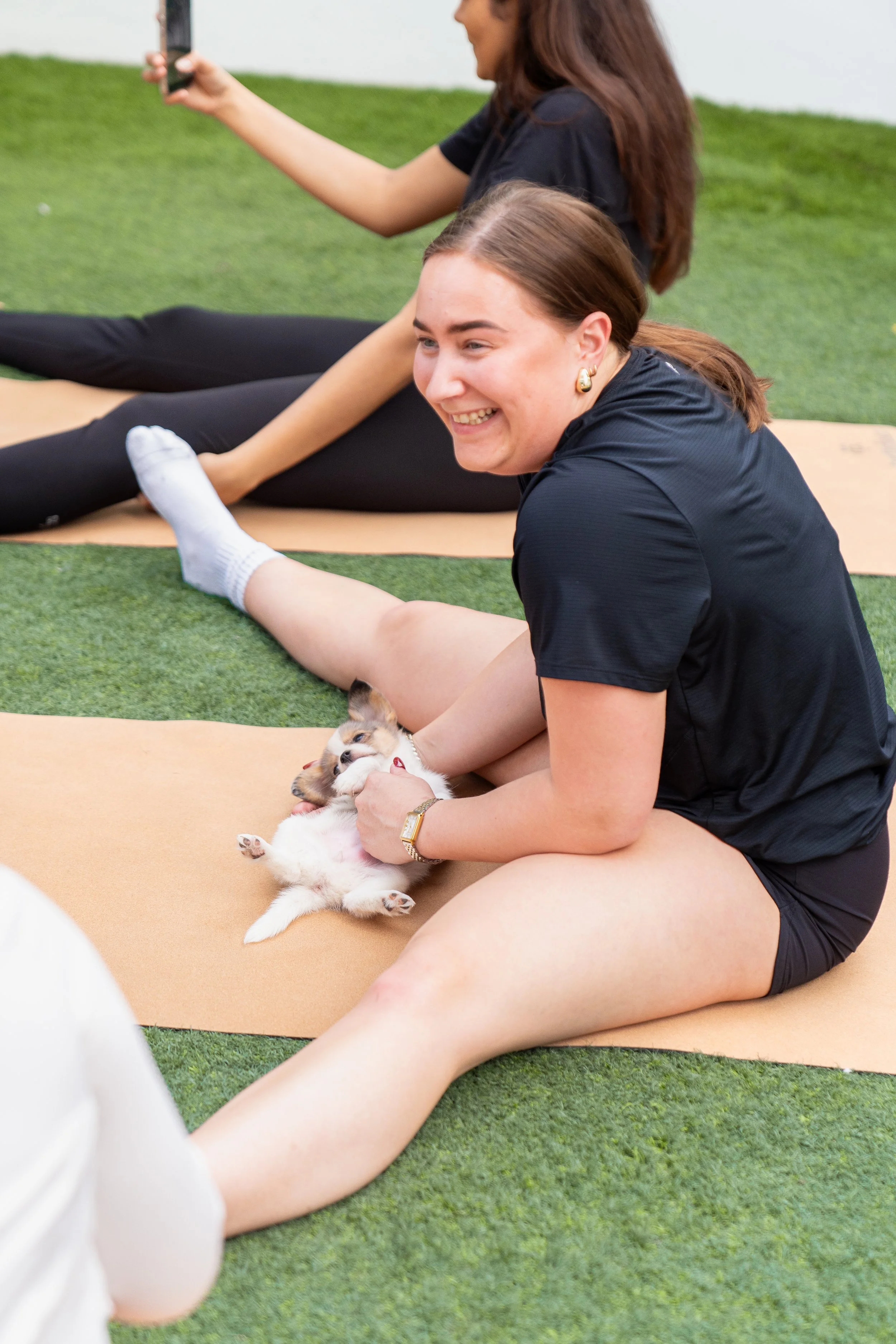 A woman in black workout clothes sitting on yoga mat, holding a cute puppy, while smiling. Other women doing stretches on yoga mats nearby.