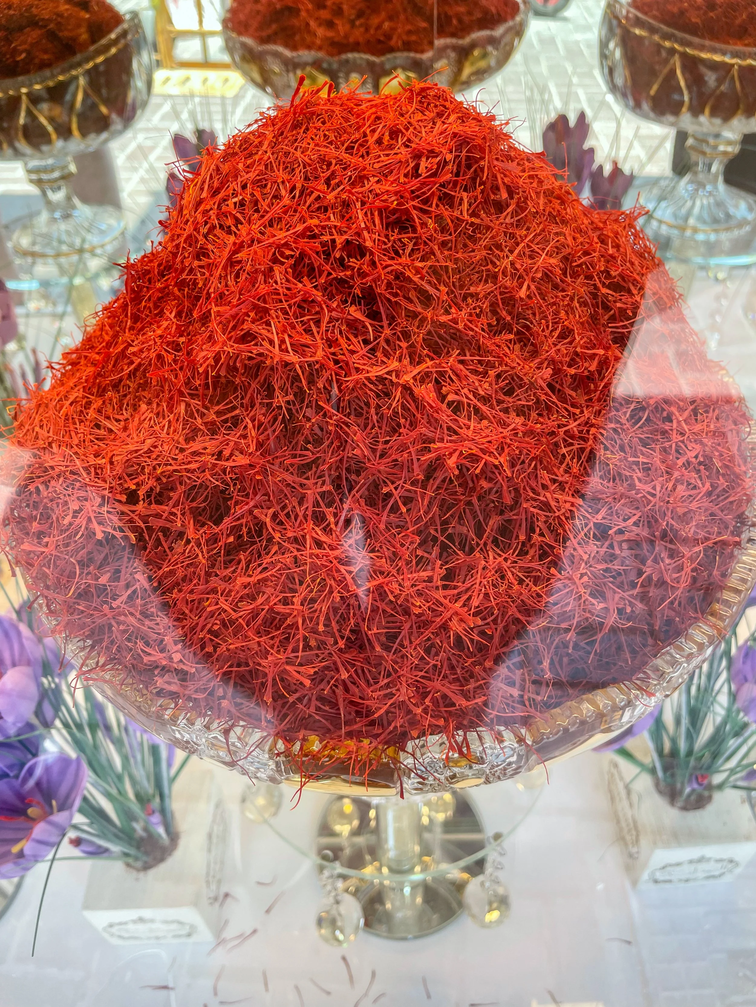 A glass display stand holding a mound of vibrant red saffron threads, with purple flowers and other glass containers in the background.
