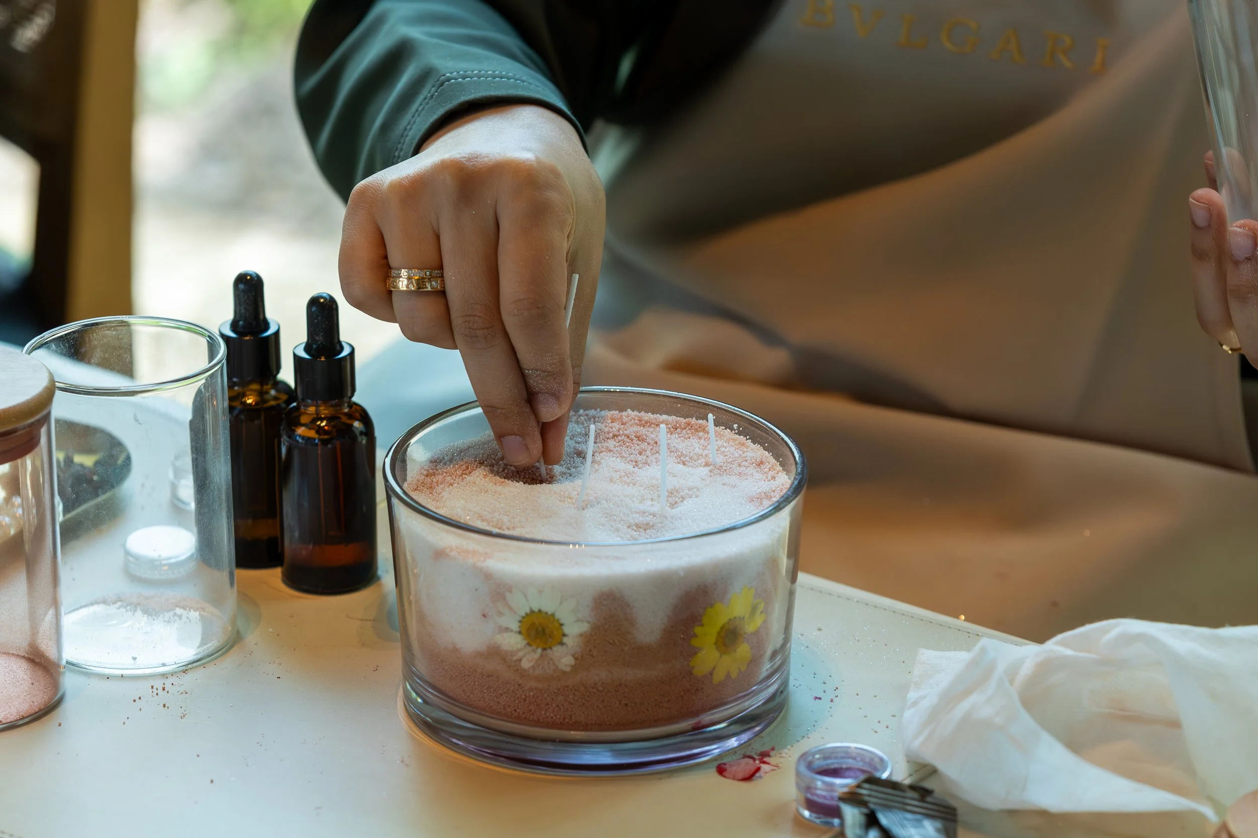 Person decorates a candle with salt and essential oils, surrounded by small bottles and candles.