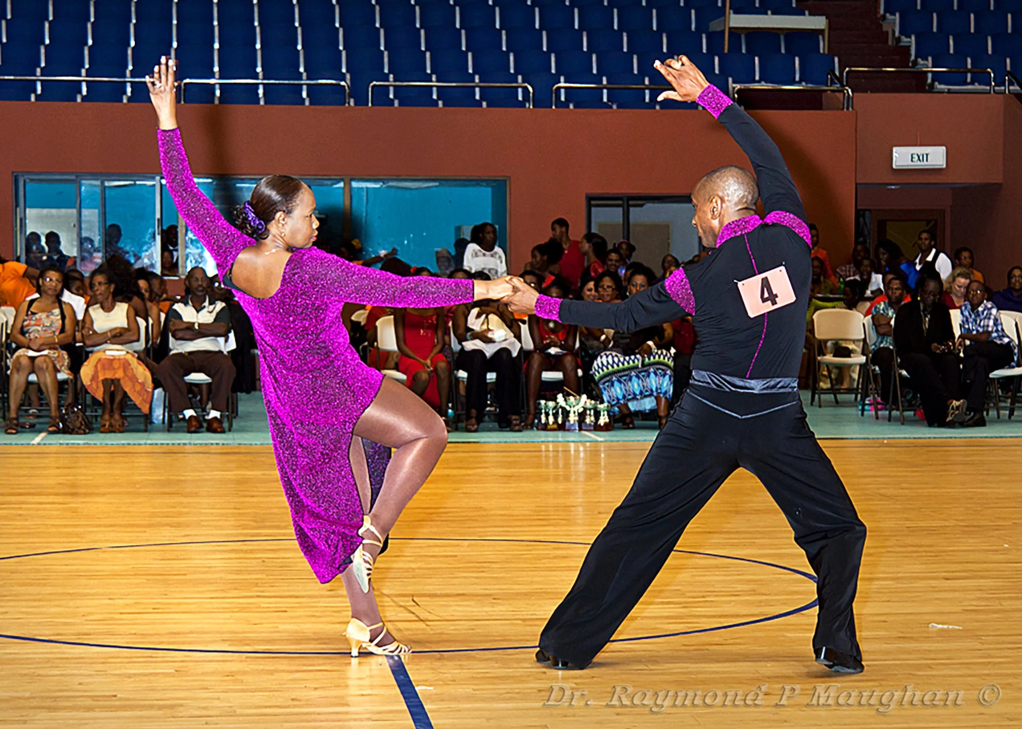 A couple dancing ballroom, with the woman in a purple dress and the man in black, surrounded by an audience seated in chairs, in an indoor hall.