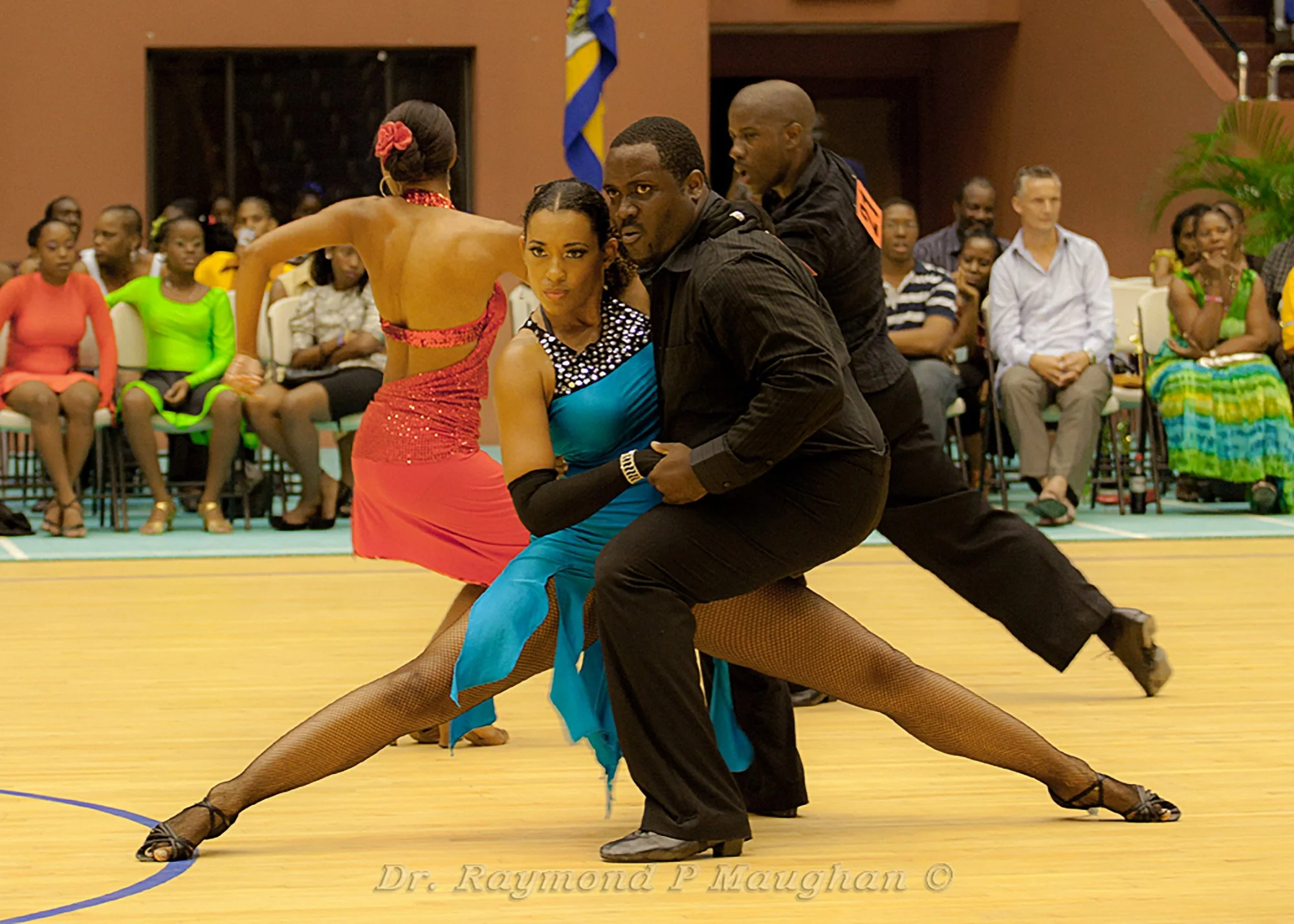 A ballroom dance performance with a male and female dancer in colorful costumes, striking a pose on the dance floor, while an audience watches in the background.