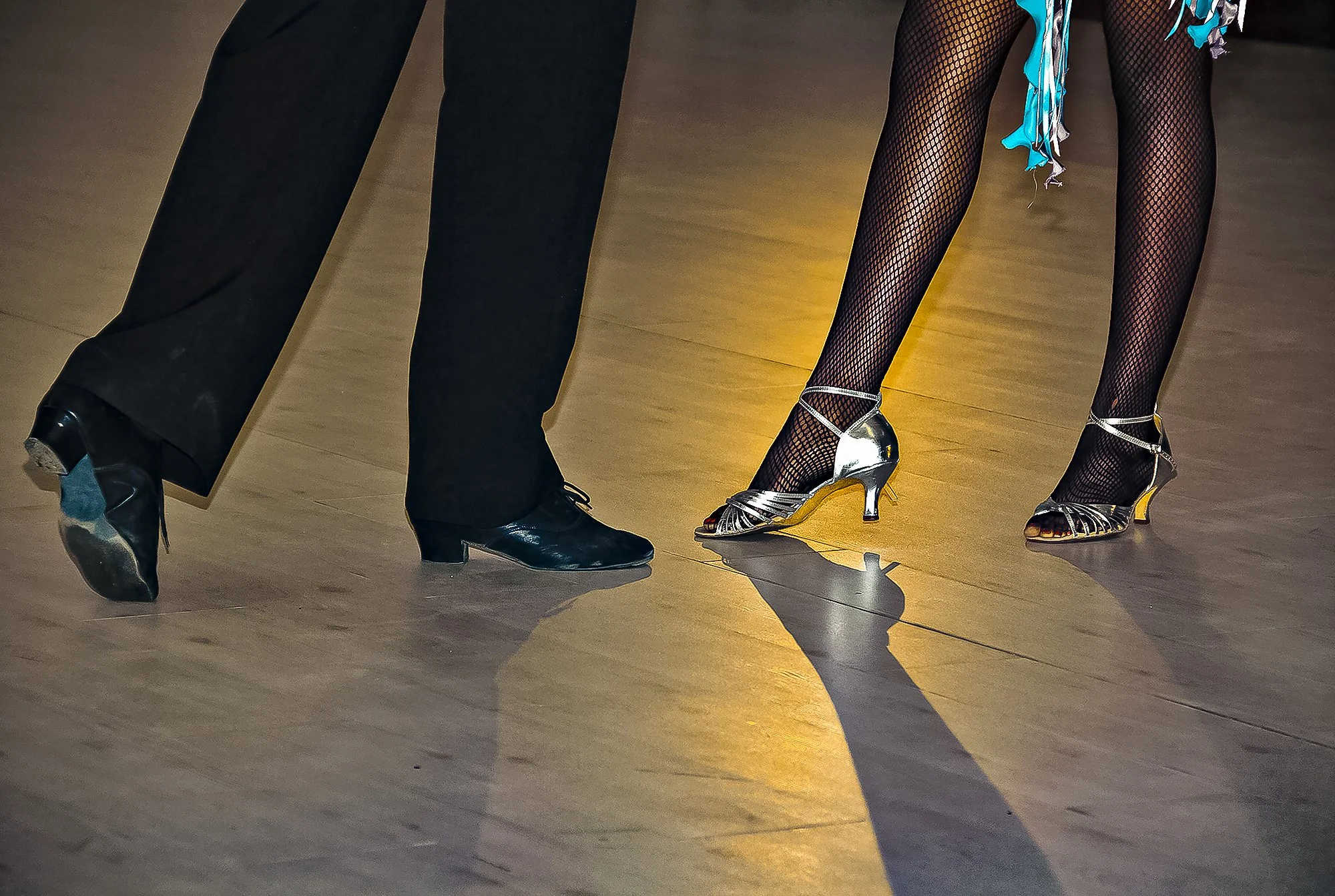 Close-up of the legs and feet of two dancers, one wearing black pants and shoes, the other wearing fishnet stockings and high heels, dancing on a wooden floor with a warm light reflecting on the surface.
