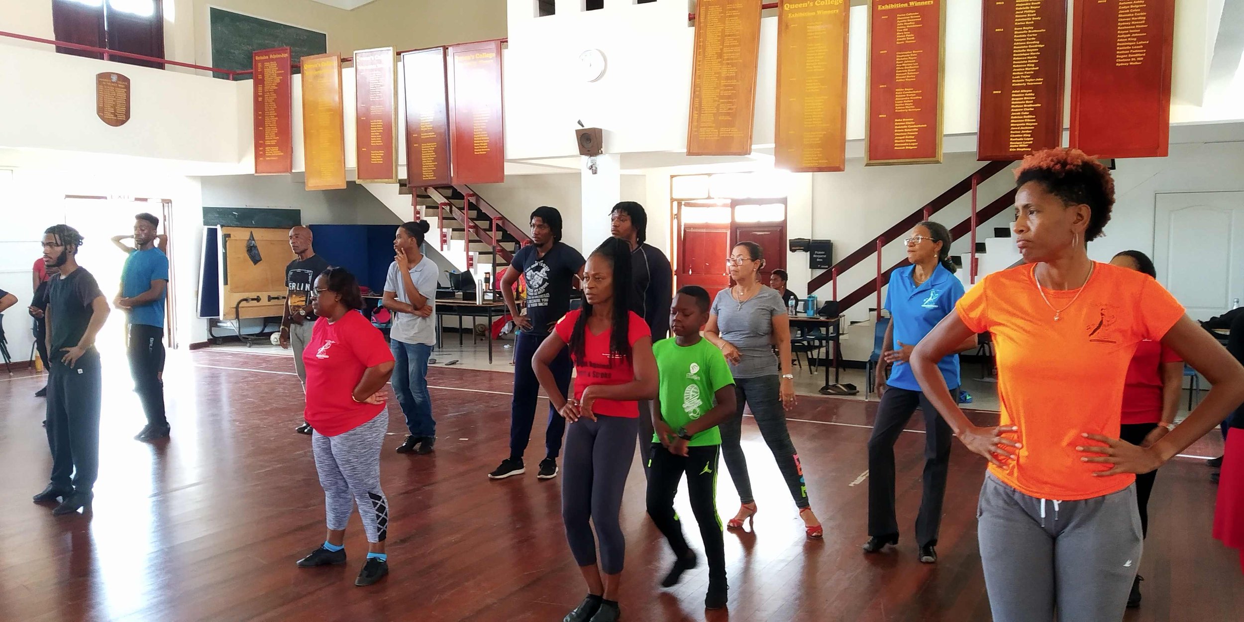 People participating in a group exercise or dance class inside a gymnasium or community center. Some are standing with hands on hips, others with arms relaxed, all facing forward and paying attention. The space has wooden floors, ladders, and large boards with yellow text on red backgrounds hanging on the white walls.