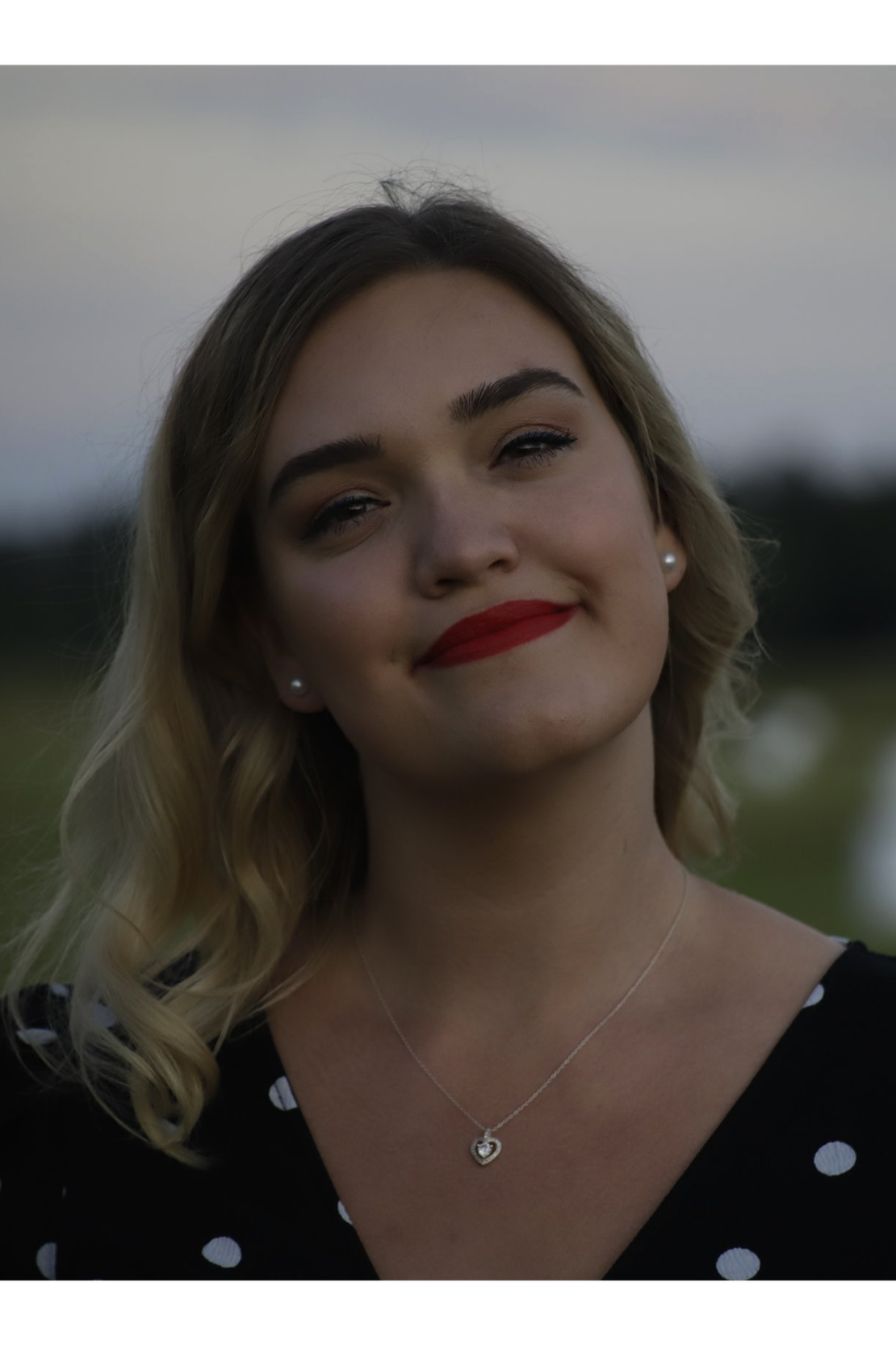A woman with blonde wavy hair, red lipstick, and pearl earrings smiling, wearing a black dress with white polka dots and a heart-shaped necklace, outdoors during the evening or late afternoon.