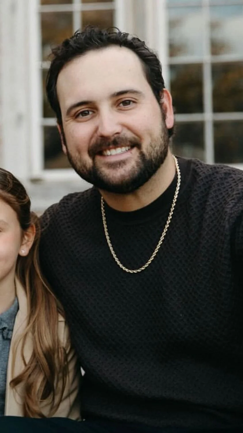 A man with short dark curly hair and a beard, smiling, wearing a black textured shirt and a gold chain, outdoors near a window.