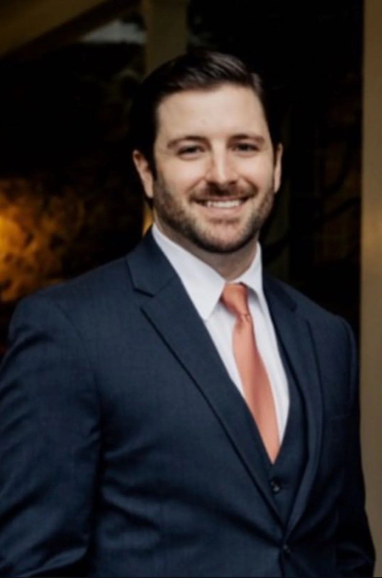 A man with dark hair and a beard, wearing a dark suit, white shirt, and peach-colored tie, smiling at the camera.