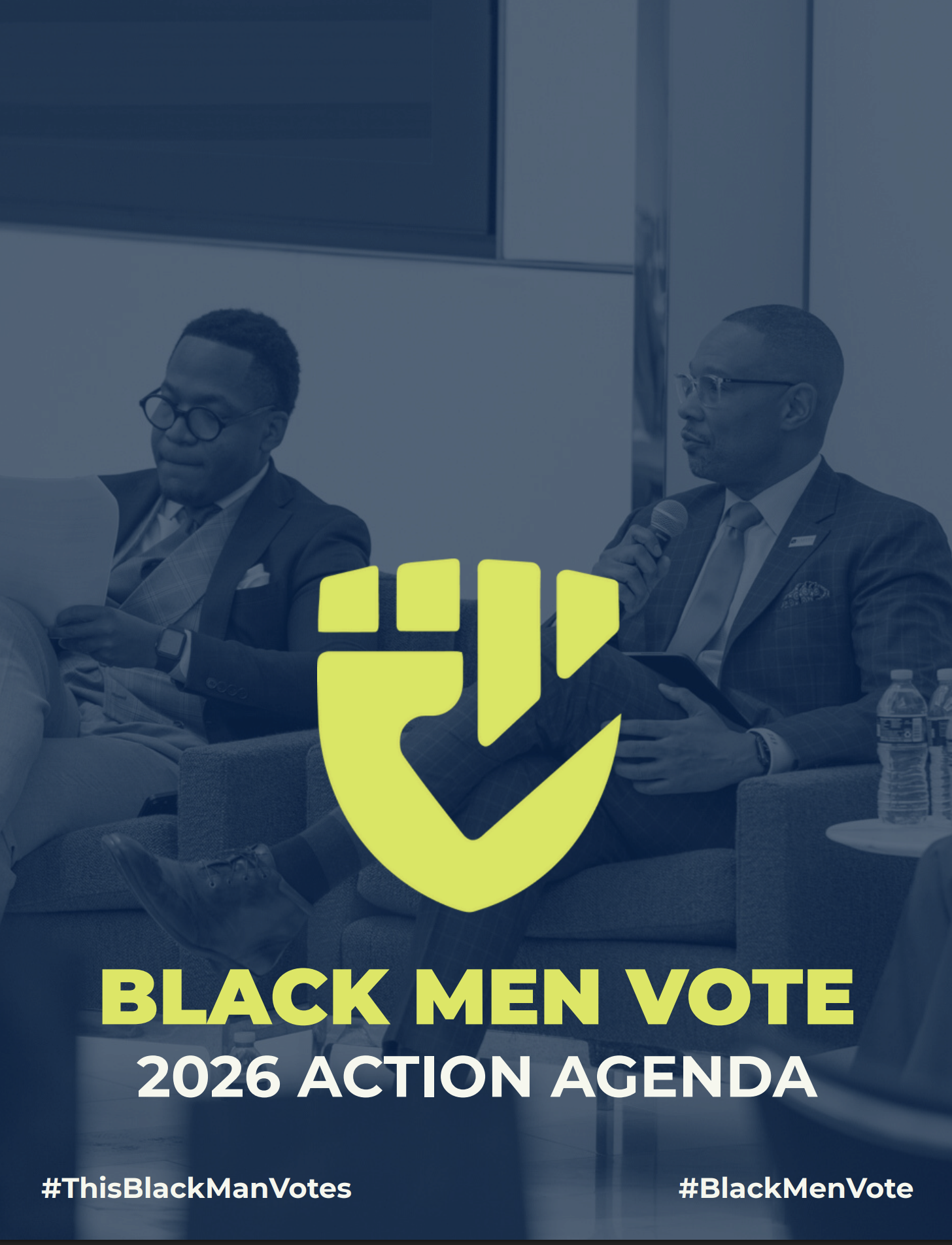 Two Black men in business suits sitting on stage, one holding a microphone, during a panel discussion at an event promoting Black men's voting rights and policies for 2026.