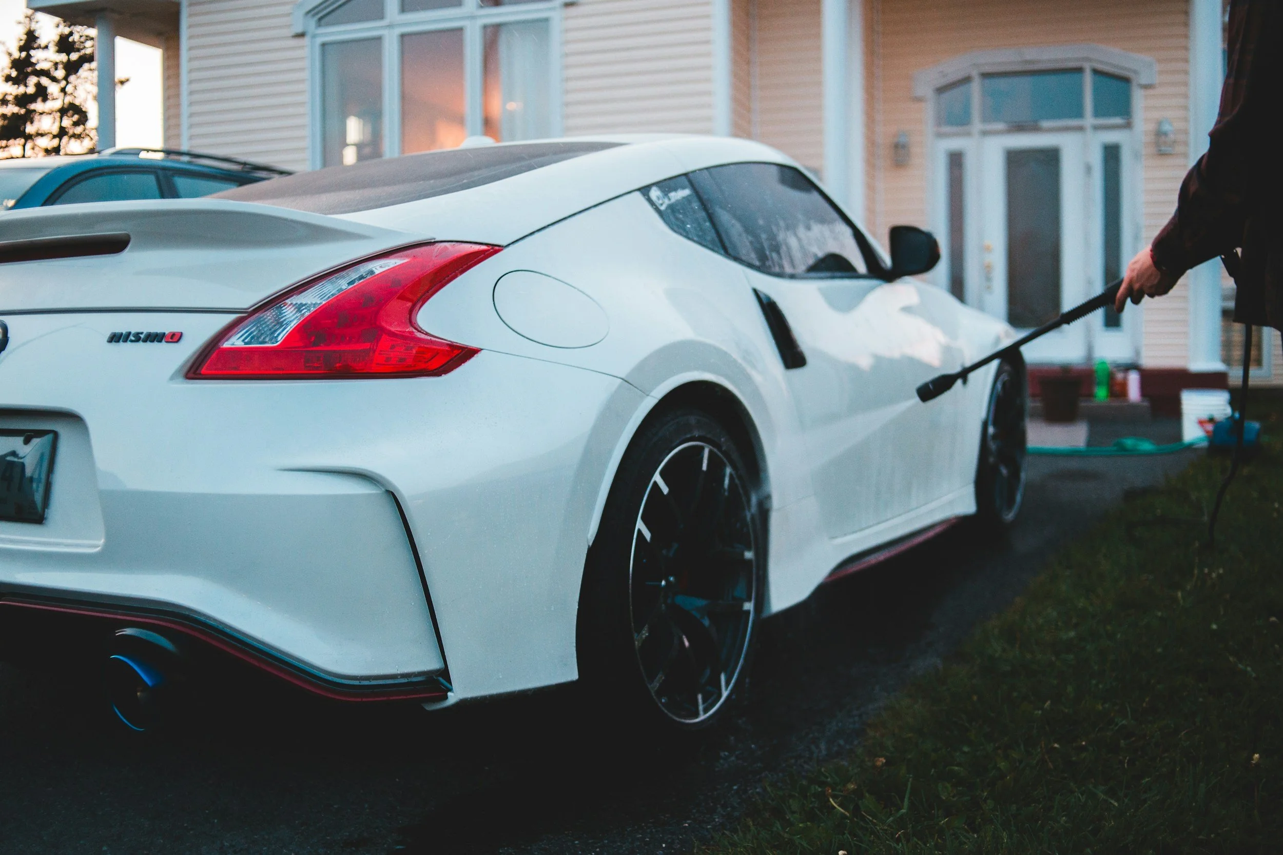 Person washing a white Nissan 370Z sports car with a hose outside a house during sunset.