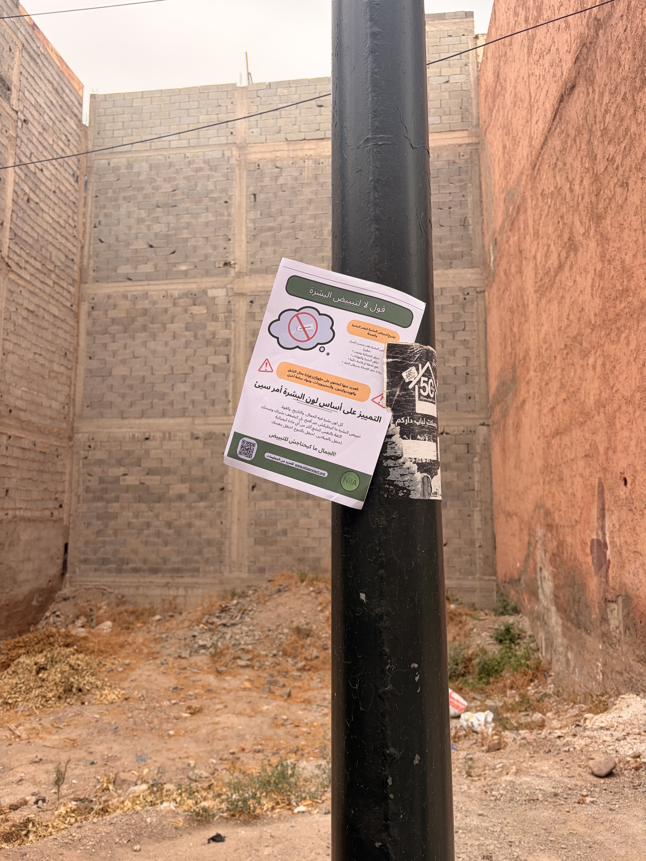 A black pole with a skin health flyer attached to it in an outdoor setting with dirt ground, scattered trash, and unfinished brick walls in the background.