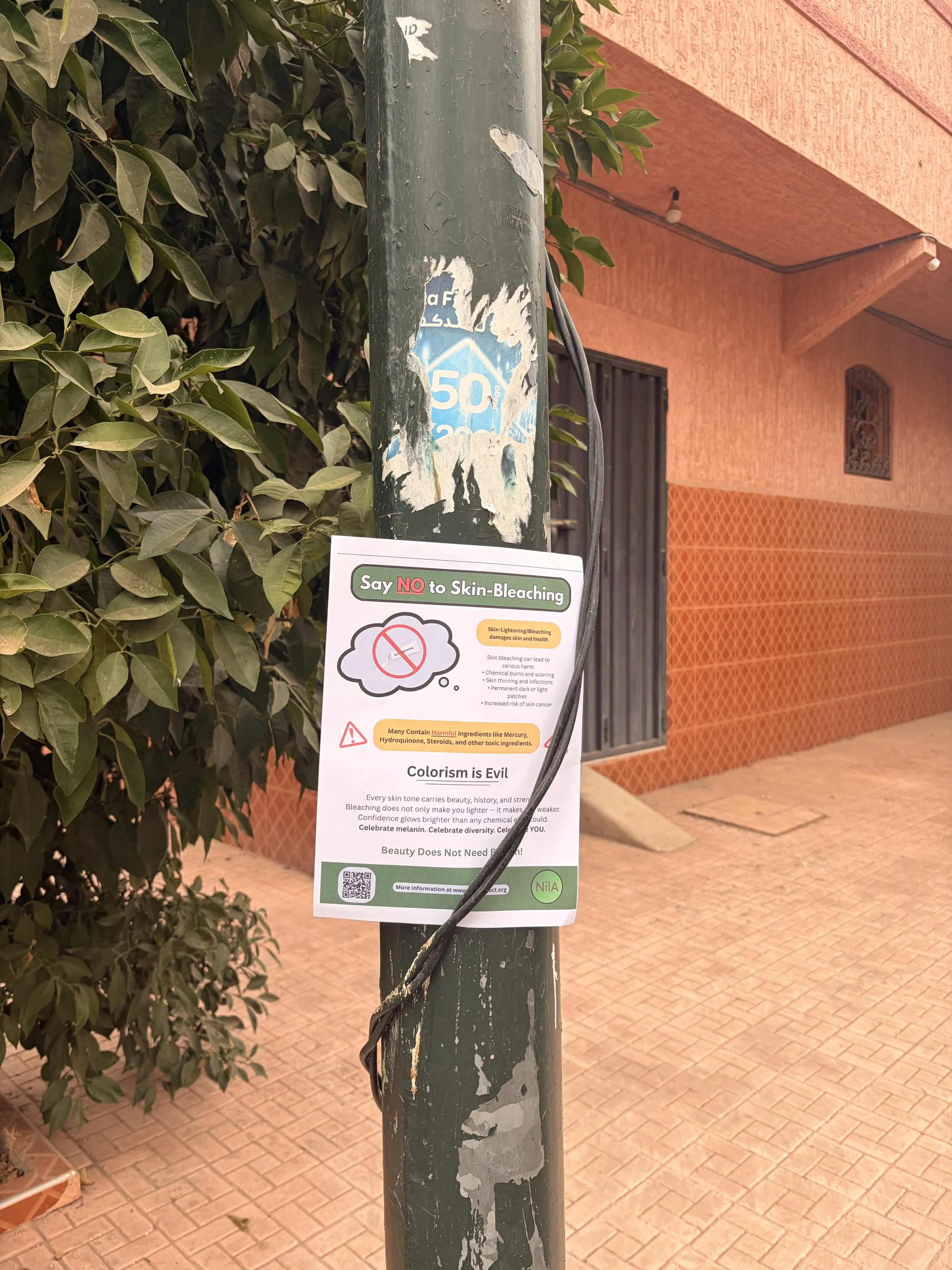 A metal signpost with a skin health flyer attached, located on a sidewalk near some greenery and a building with orange-brown textured walls and a tiled surface.