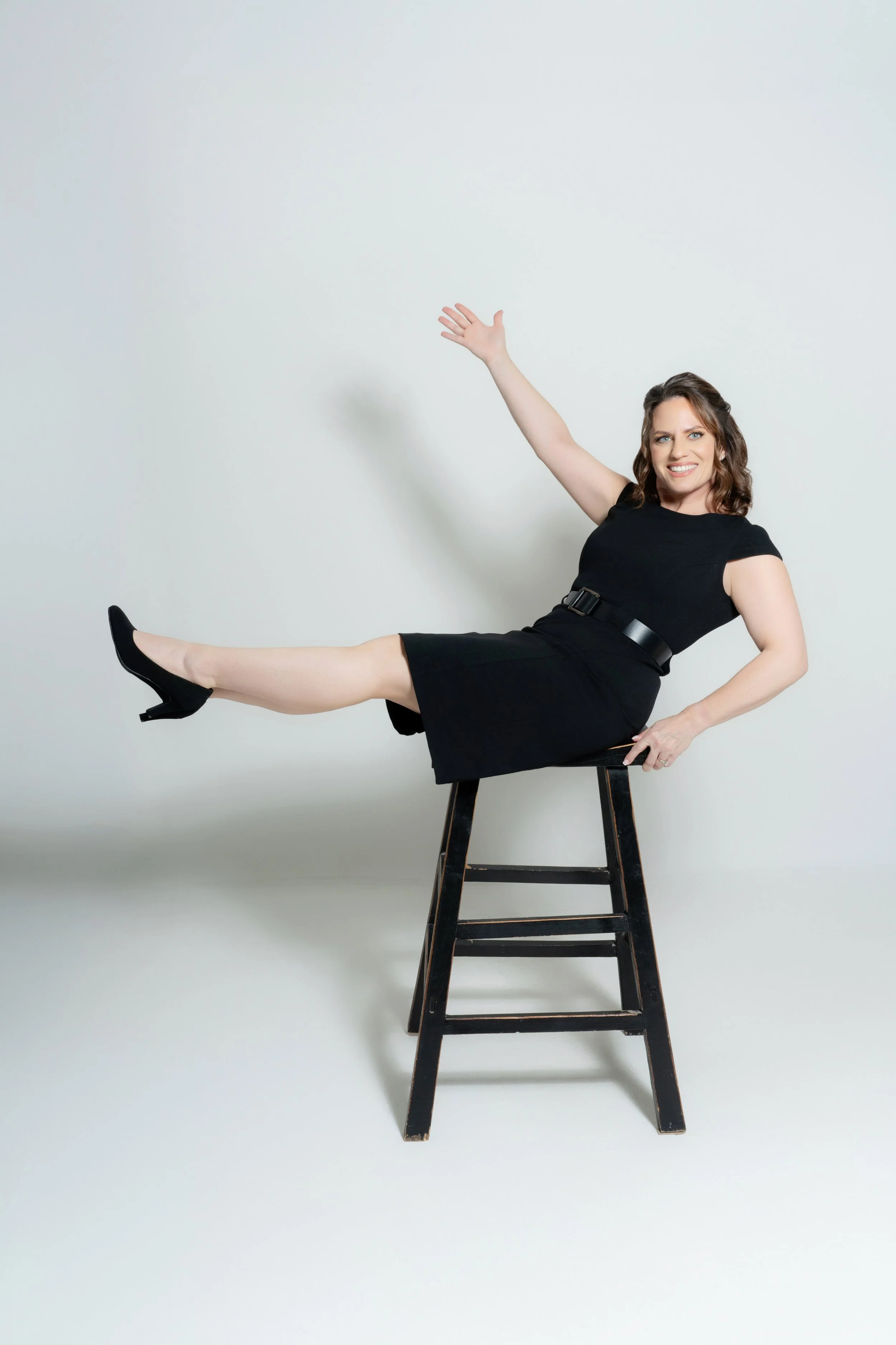 A woman sitting sideways on a black chair, smiling, with one arm raised and the other resting on her lap, dressed in a black dress and black heels, against a plain white background.