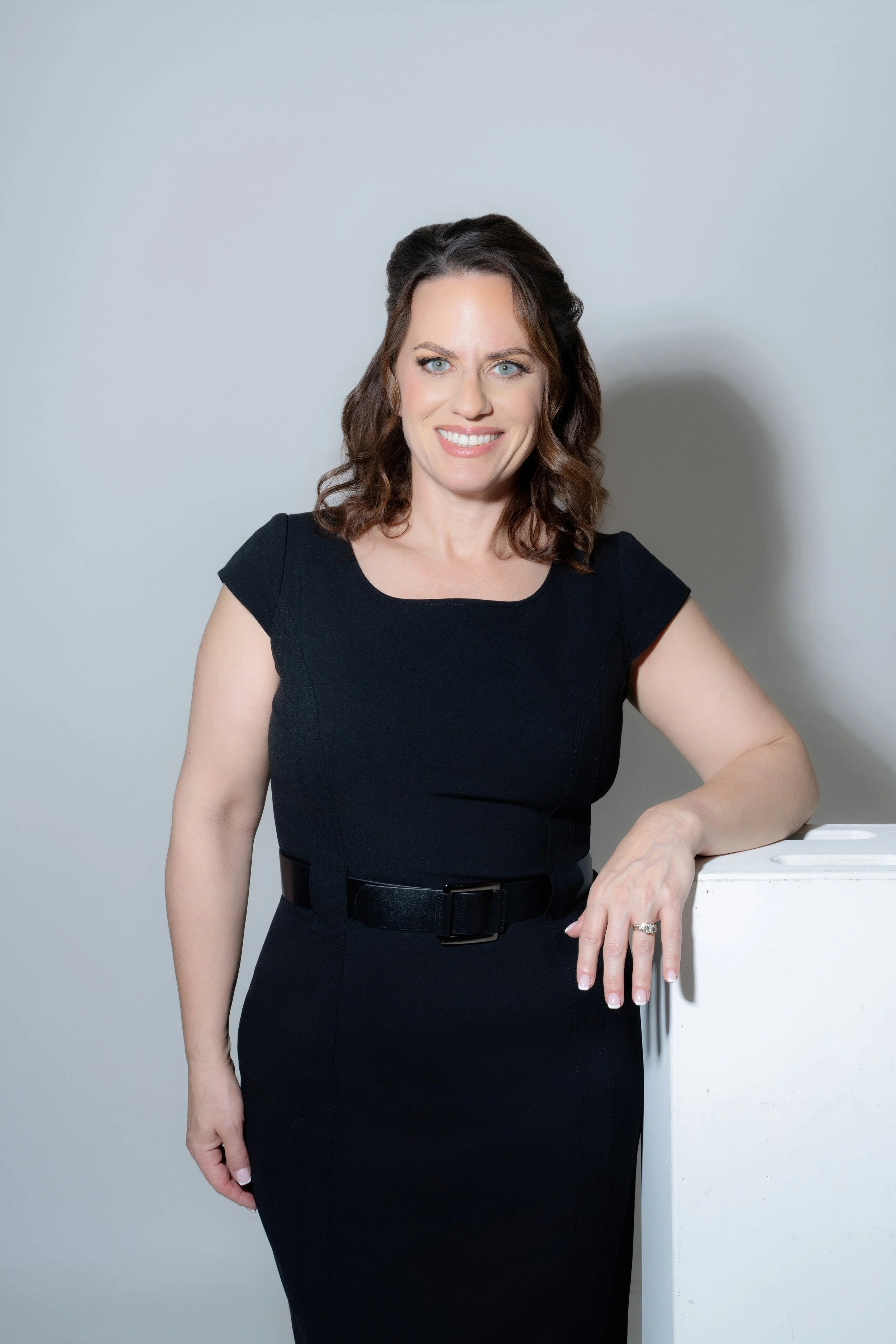 A woman with shoulder-length brown hair, wearing a black dress with a belt, standing next to a white object, smiling at the camera against a plain gray background.