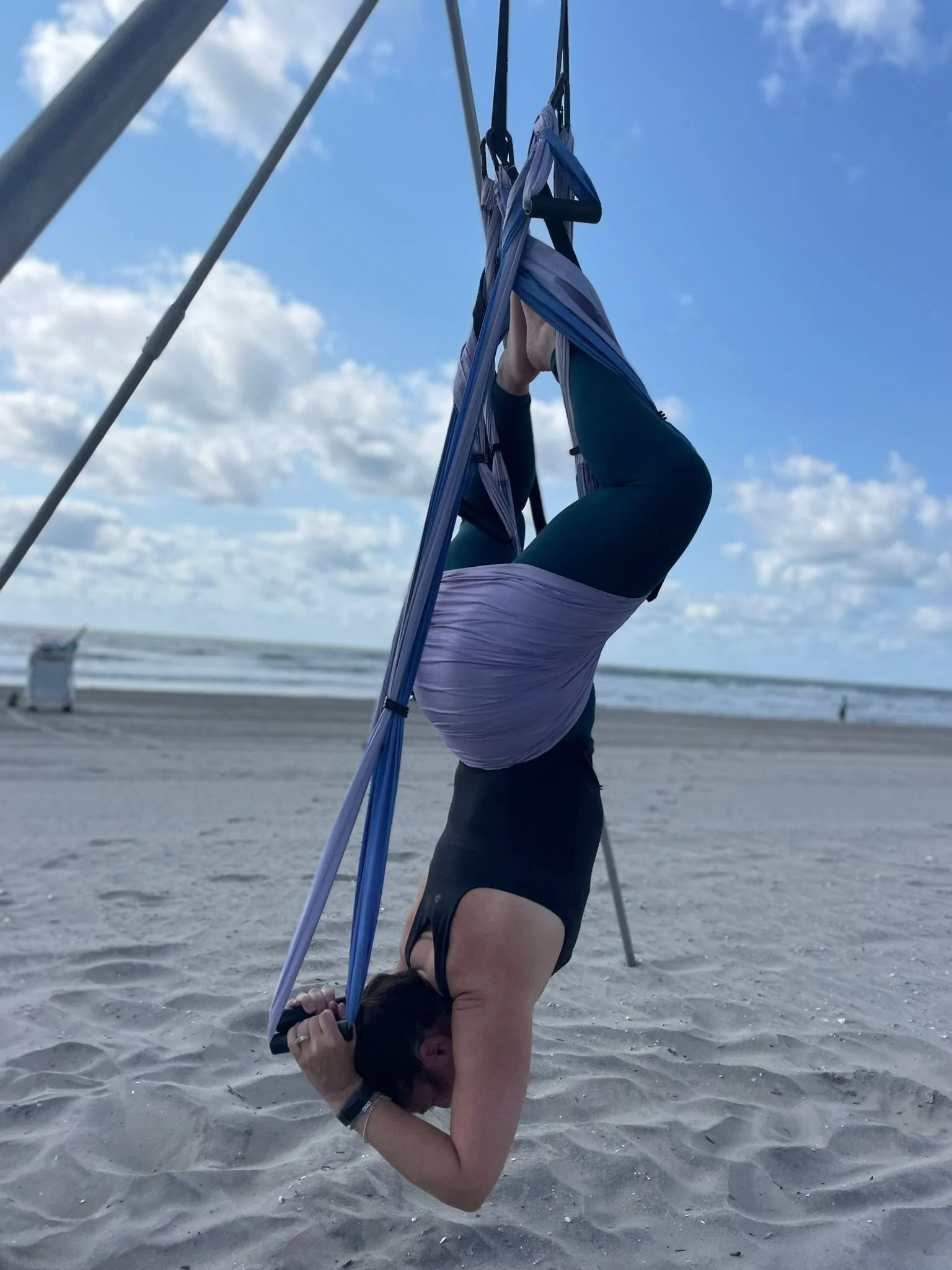 Person practicing yoga on the beach, in a headstand position with arms folded over their head, under a blue sky with clouds.