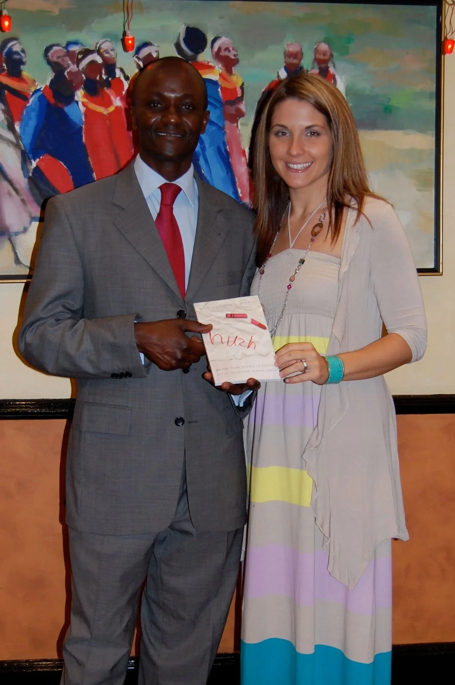 A man in a gray suit and red tie standing beside a woman in a colorful striped dress and beige cardigan, holding a check and smiling. Behind them is a colorful painting of people in traditional attire.