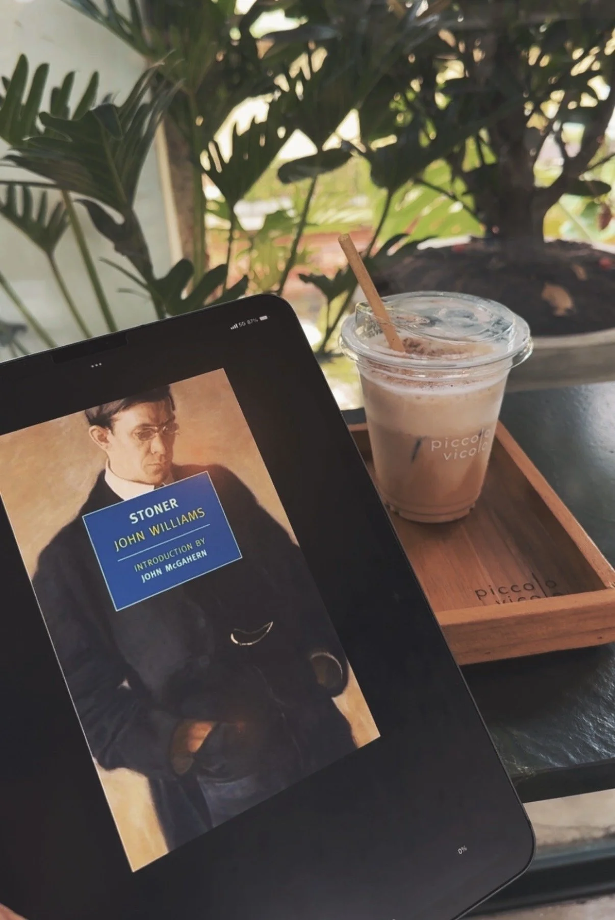 An angled shot of the book Stoner and a coffee cup showcasing a lush green background