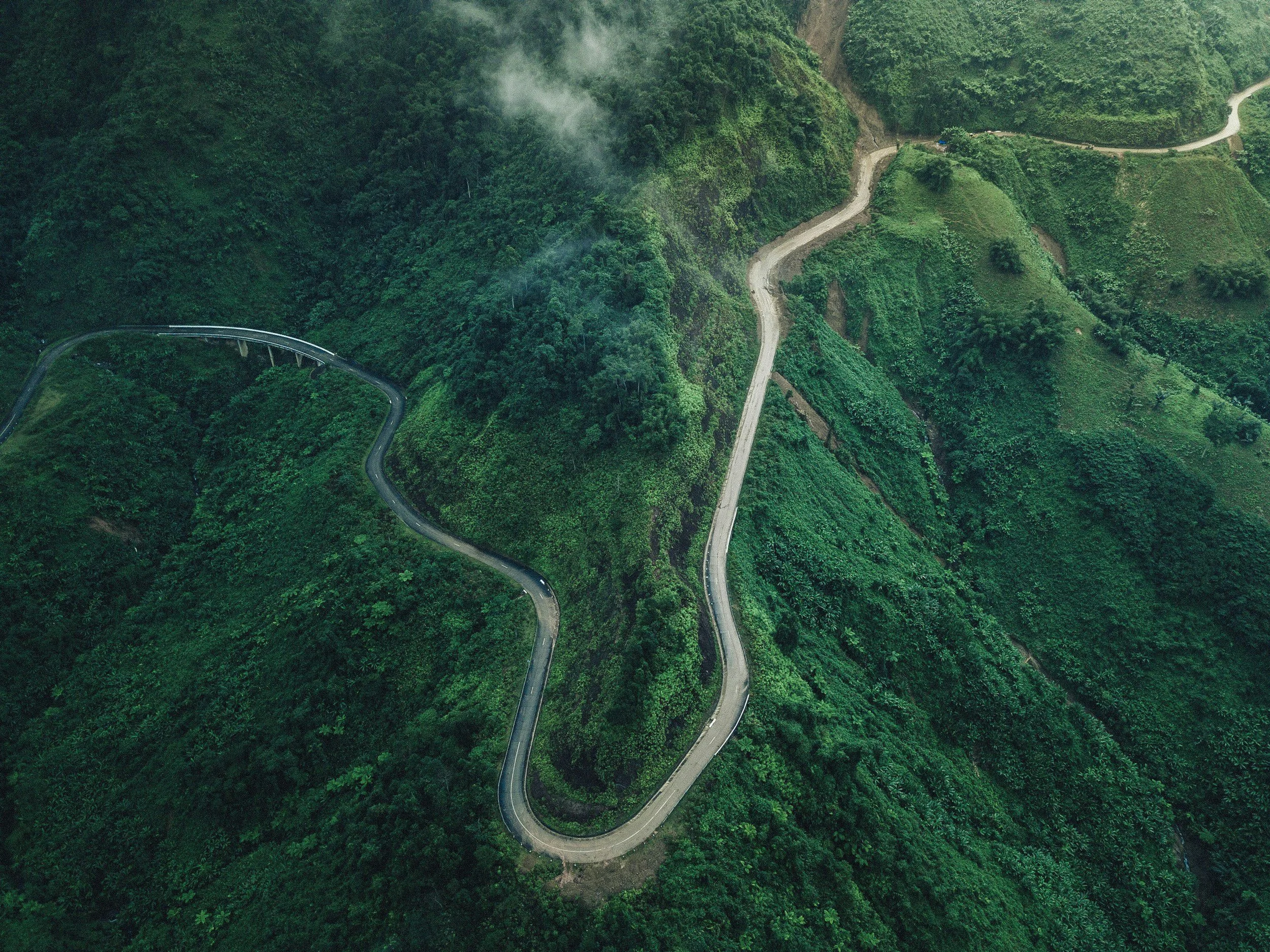 A winding mountain road through lush green hills, with a bridge on the left side and a dirt path crossing the terrain.