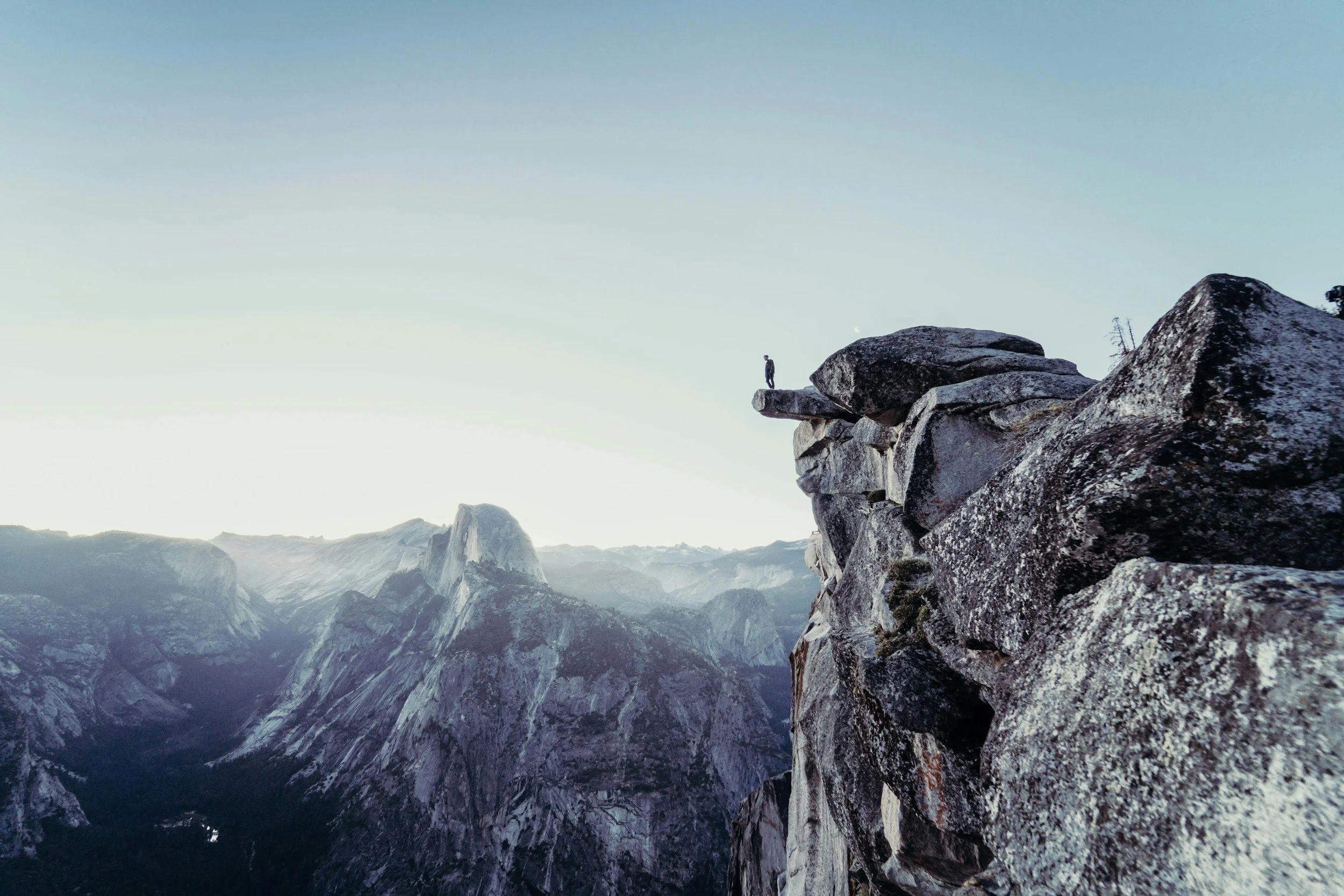 A person standing on a small ledge at the end of a rocky cliff, overlooking a mountain valley with a prominent mountain peak in the background during daylight.