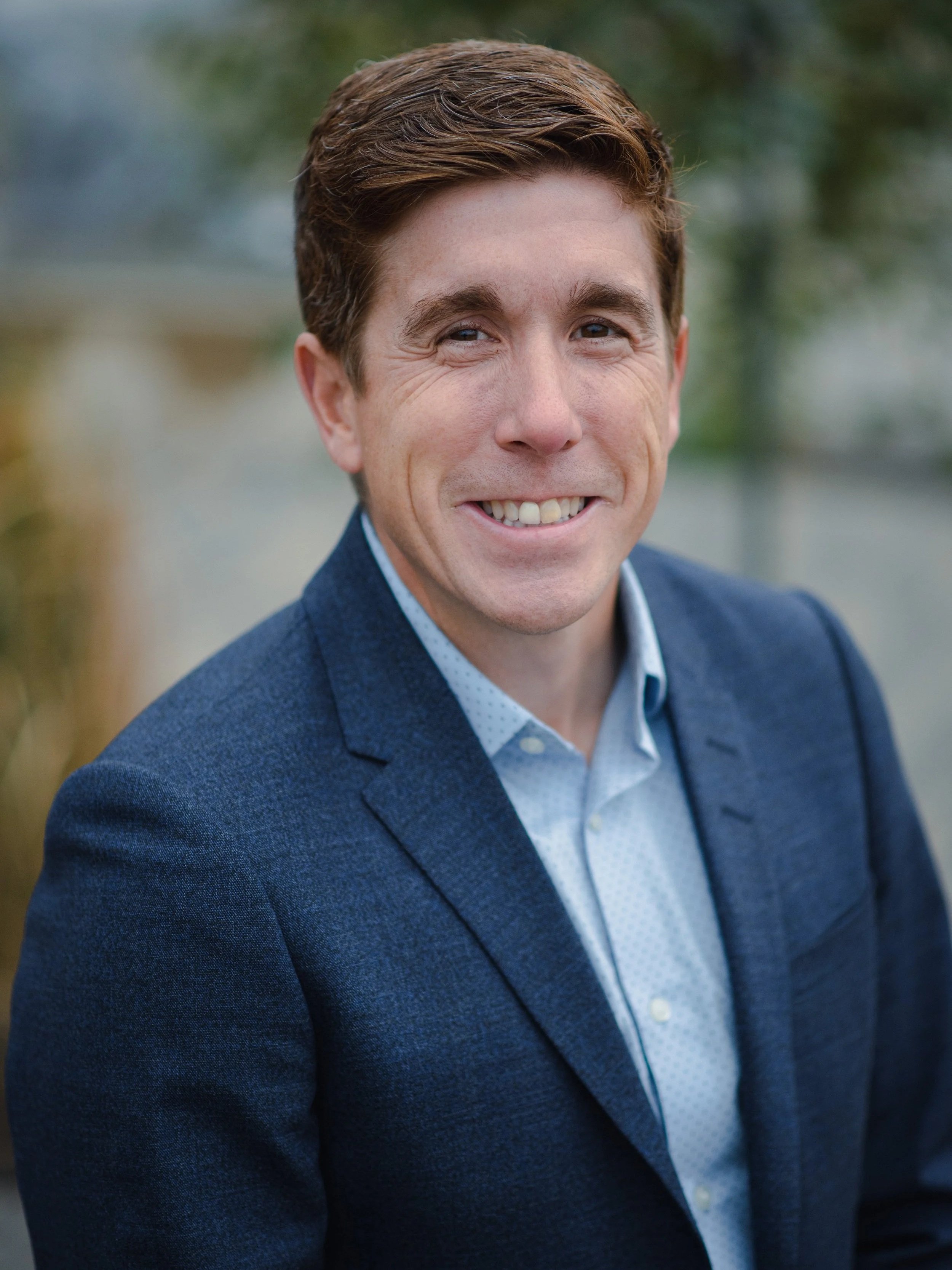 A man with brown hair wearing a blue blazer and white shirt smiling outdoors.