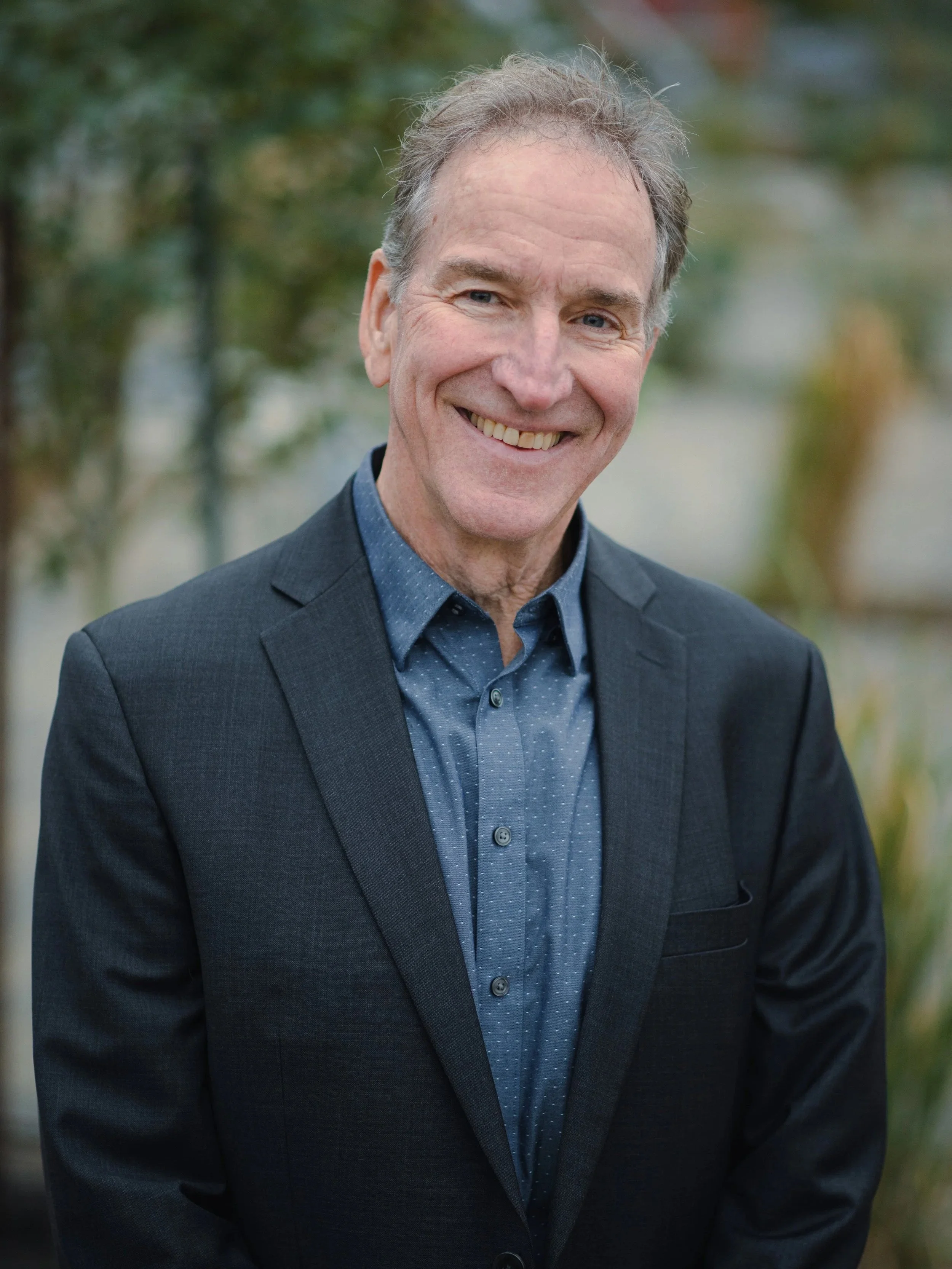 A smiling middle-aged man wearing a dark suit jacket and blue button-up shirt, standing outdoors with blurred trees and foliage in the background.