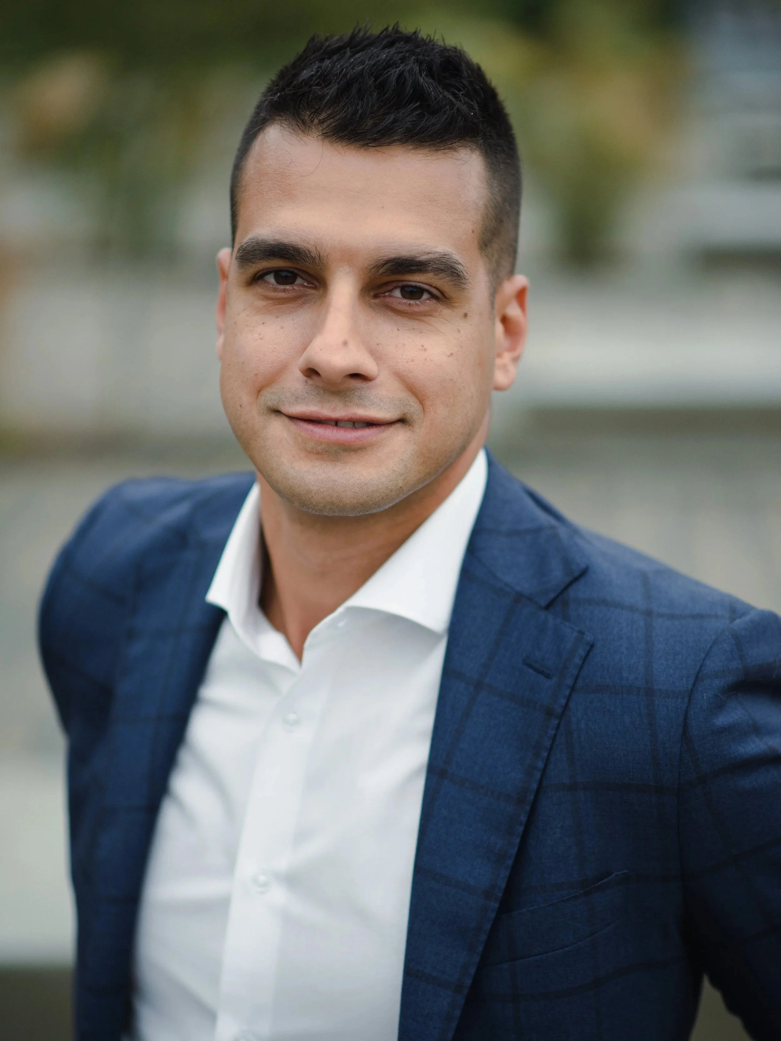 Close-up portrait of a man in a navy blazer and white shirt outdoors, smiling confidently.